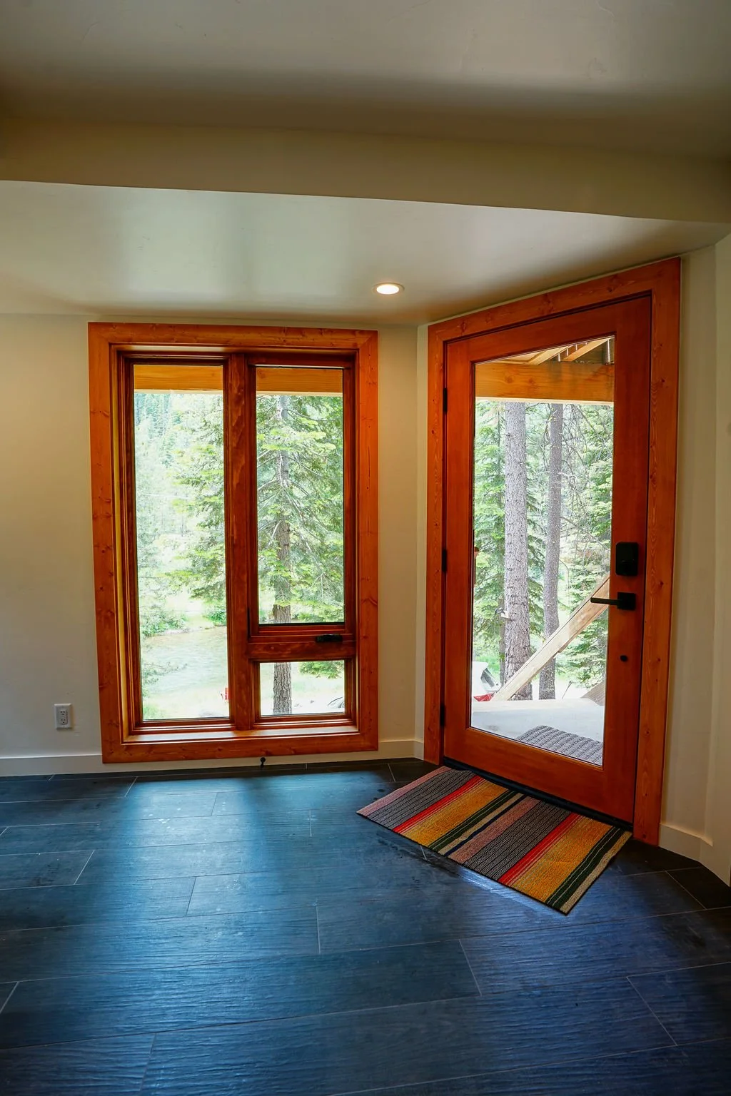 Bright entryway with a red front door with glass panel, tall windows, dark wood flooring, white walls, and a colorful rainbow rug.