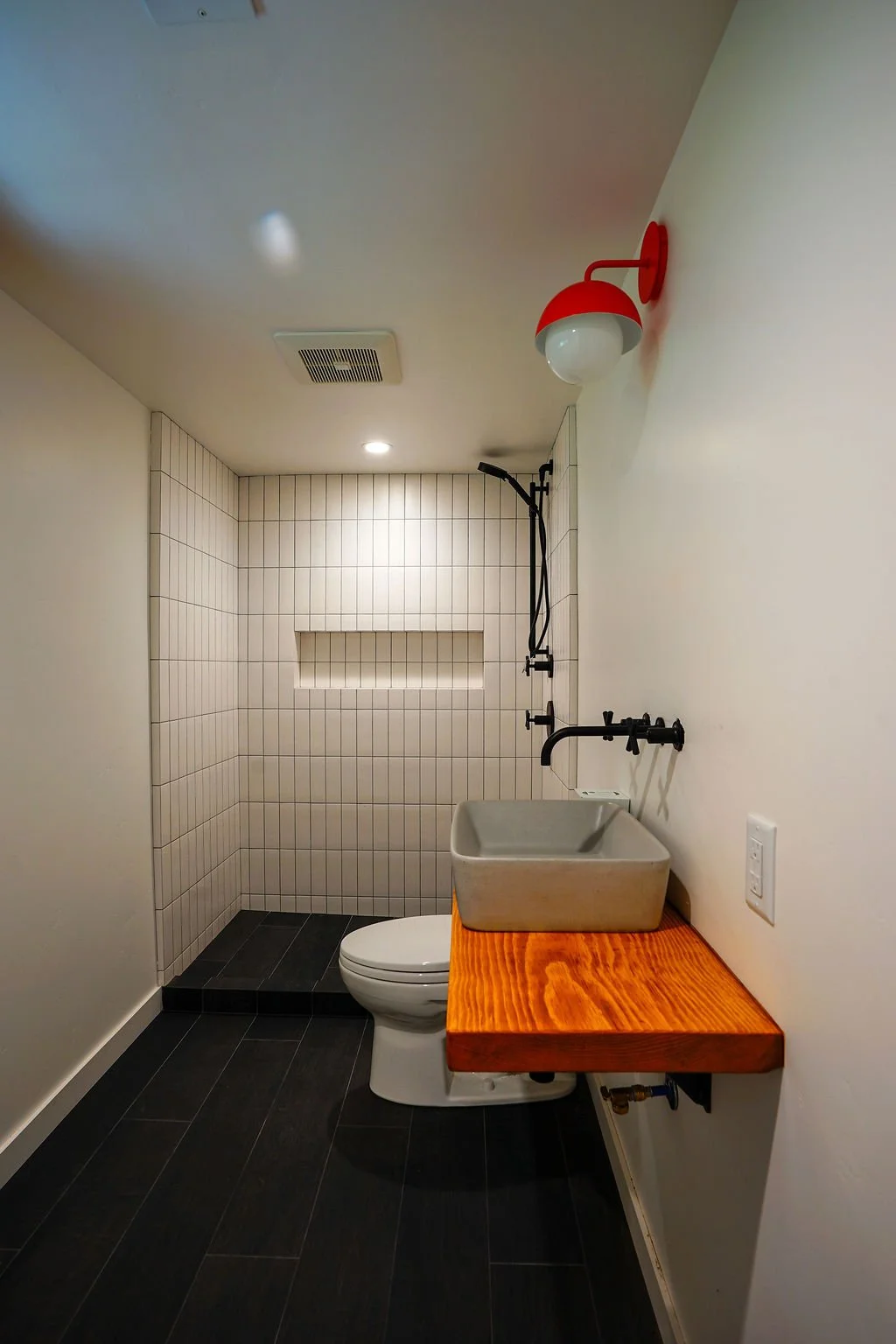 Narrow bathroom with white walls, a white tiled shower, black flooring and fixtures, a floating wood vanity with gray sink basin, and a small red wall light.