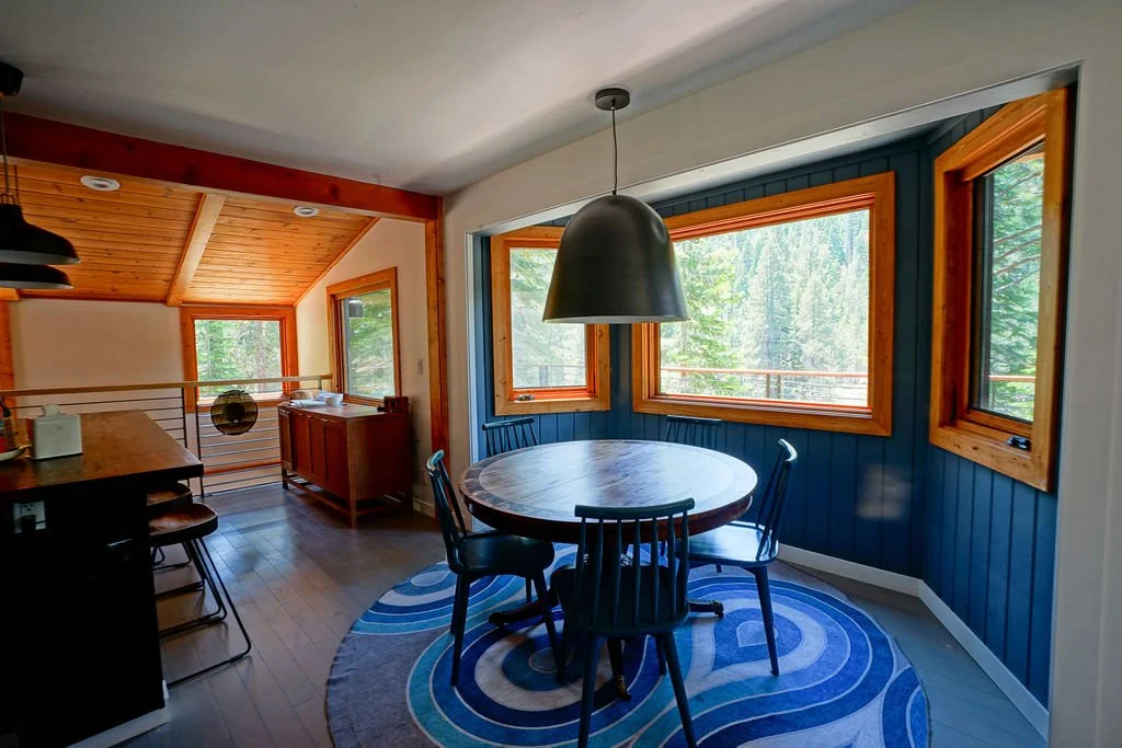 Bright dining area with circular table and blue wooden chairs and large green pendant light with large windows set into a popped out blue accent wall and an open view into the kitchen.