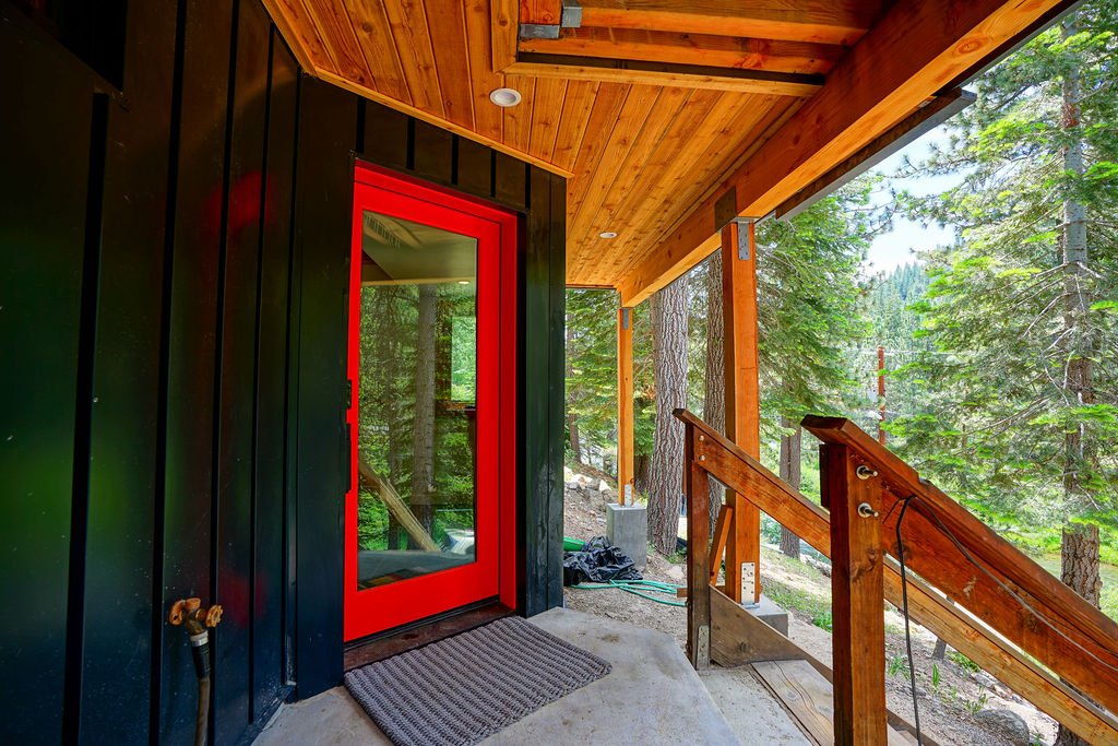 Small cabin-style home with dark gray siding, wood railings, a red front door with a large glass panel, and a wood deck surrounded by pine trees.