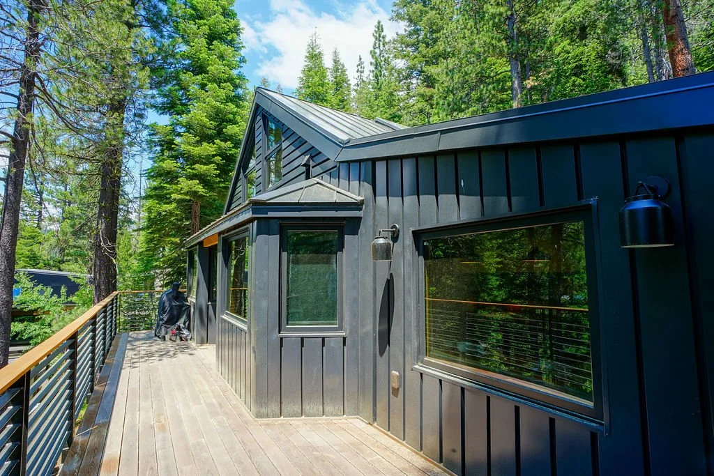 Black-sided cabin-style home with a peaked roof, large wood deck, modern metal railing, and a wooded natural setting.