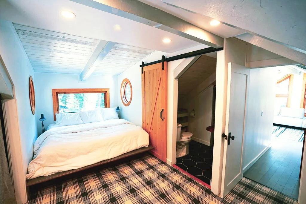 Loft bedroom with blue and white plaid carpet, a queen bed in a cozy nook with small windows, and a barn door leading to a half bathroom.