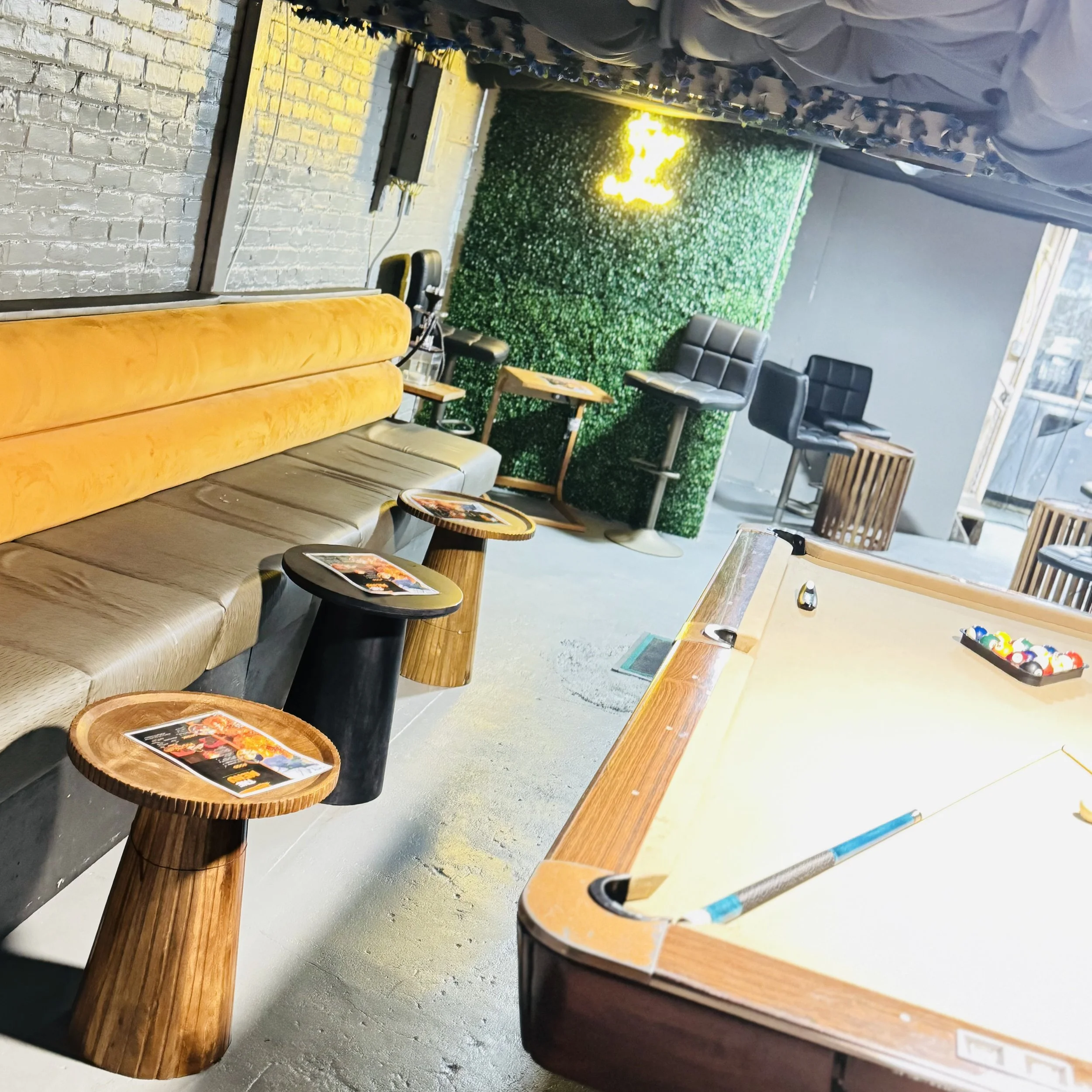 Interior of a modern recreational space featuring a pool table with billiard balls arranged for play. There is seating with yellow cushioned benches, small round wooden tables with mats, and high chairs near a green wall. A ceiling with fabric drapes