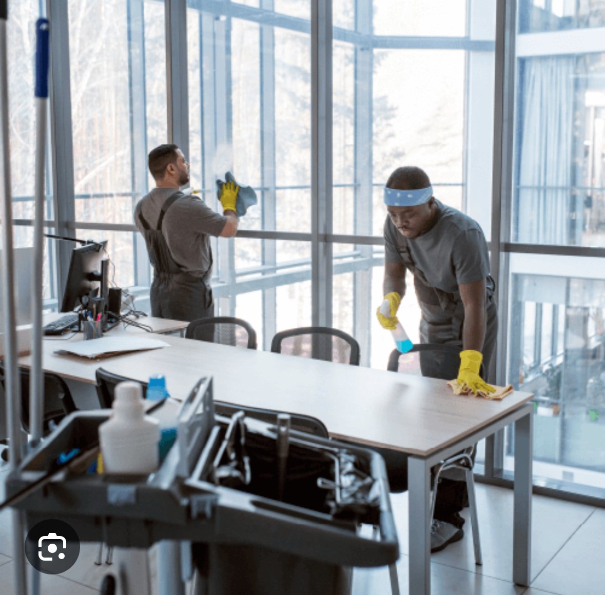 Two janitors cleaning an office space; one wiping a window, another cleaning a table. Cleaning cart with supplies is visible in the foreground.