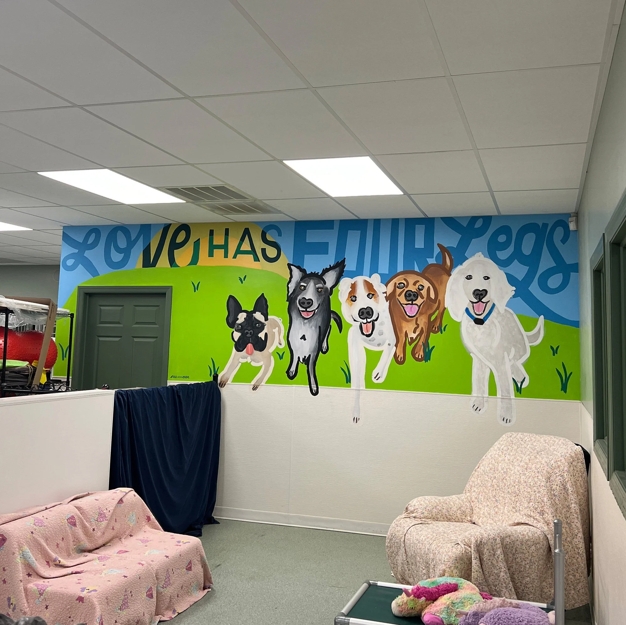 Colorful wall mural of five dogs running on green grass with a blue sky background, with partially visible words 'Love HAS Four Legs' The room has a pink blanket on a small couch, an armchair, and some toys on a small table.