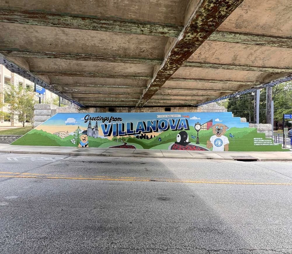 Colorful mural on a bridge underpass featuring the words 'Greetings from Villanova' and cartoon images of a dog, a penguin, a clock, trees, and buildings, representing the town of Villanova.