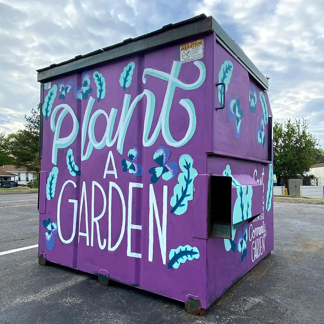 A purple dumpster with hand-painted blue and white botanical designs and the words "Plant a Garden" on the side, serving as a community gardening sales booth.
