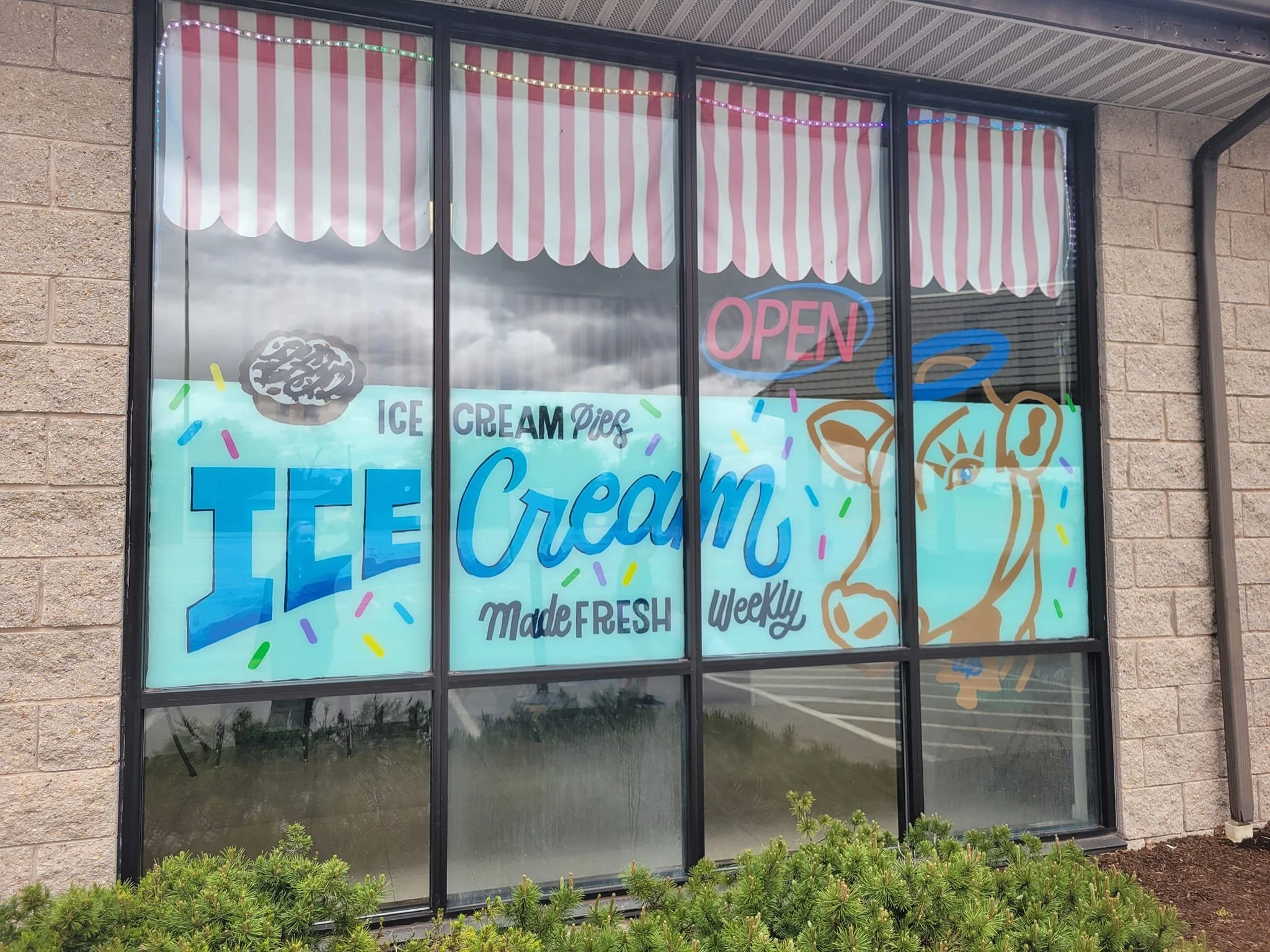 Storefront window advertising ice cream with colorful text and graphics, including a cow's face, an ice cream cone, and a face of an ice cream scoop with multicolored sprinkles, with an 'Open' sign and a red-and-white striped awning