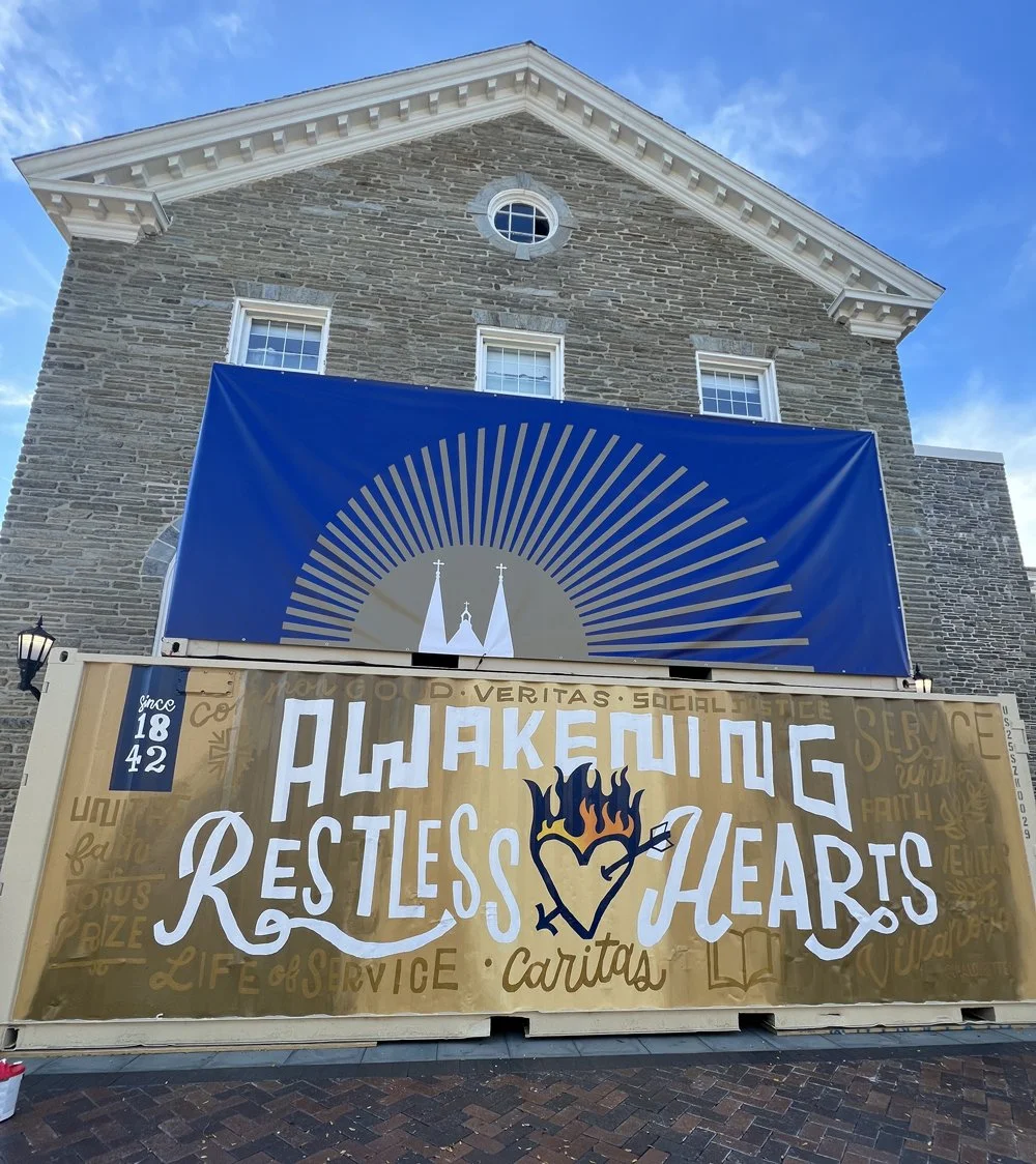 Banner on a building with the words "Awakening Restless Hearts" and a heart with a flame and a cross through it, along with the words "since 1842" and "Caritas."