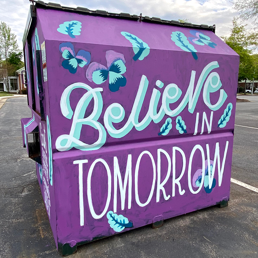 Hand-painted sign on a purple utility box with the words "Believe in Tomorrow" and decorative blue and purple butterflies and leaves.
