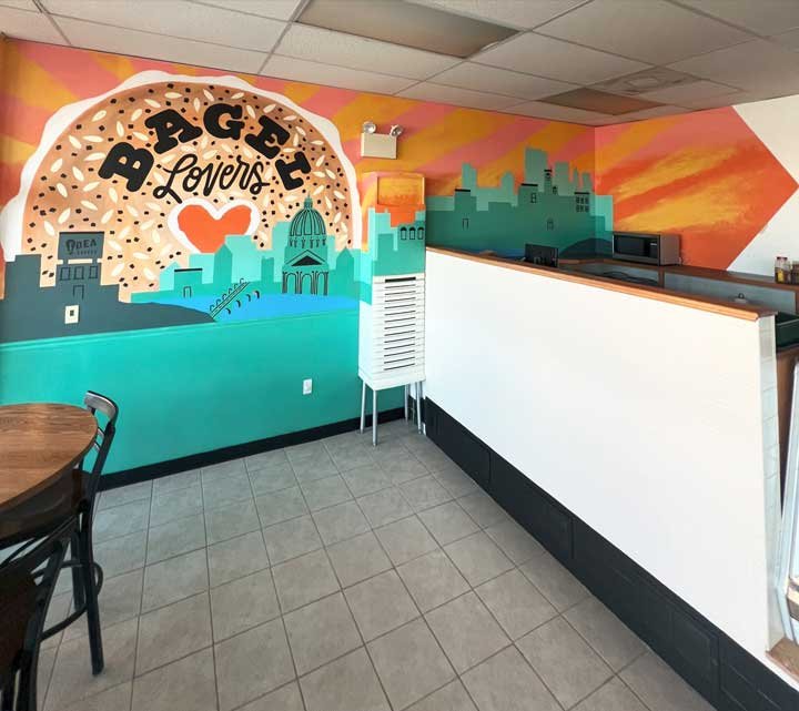 Colorful mural on the wall advertising Bagel Lover's with a sunset sky, city skyline, and a large sandwich with toppings underneath the restaurant's name.