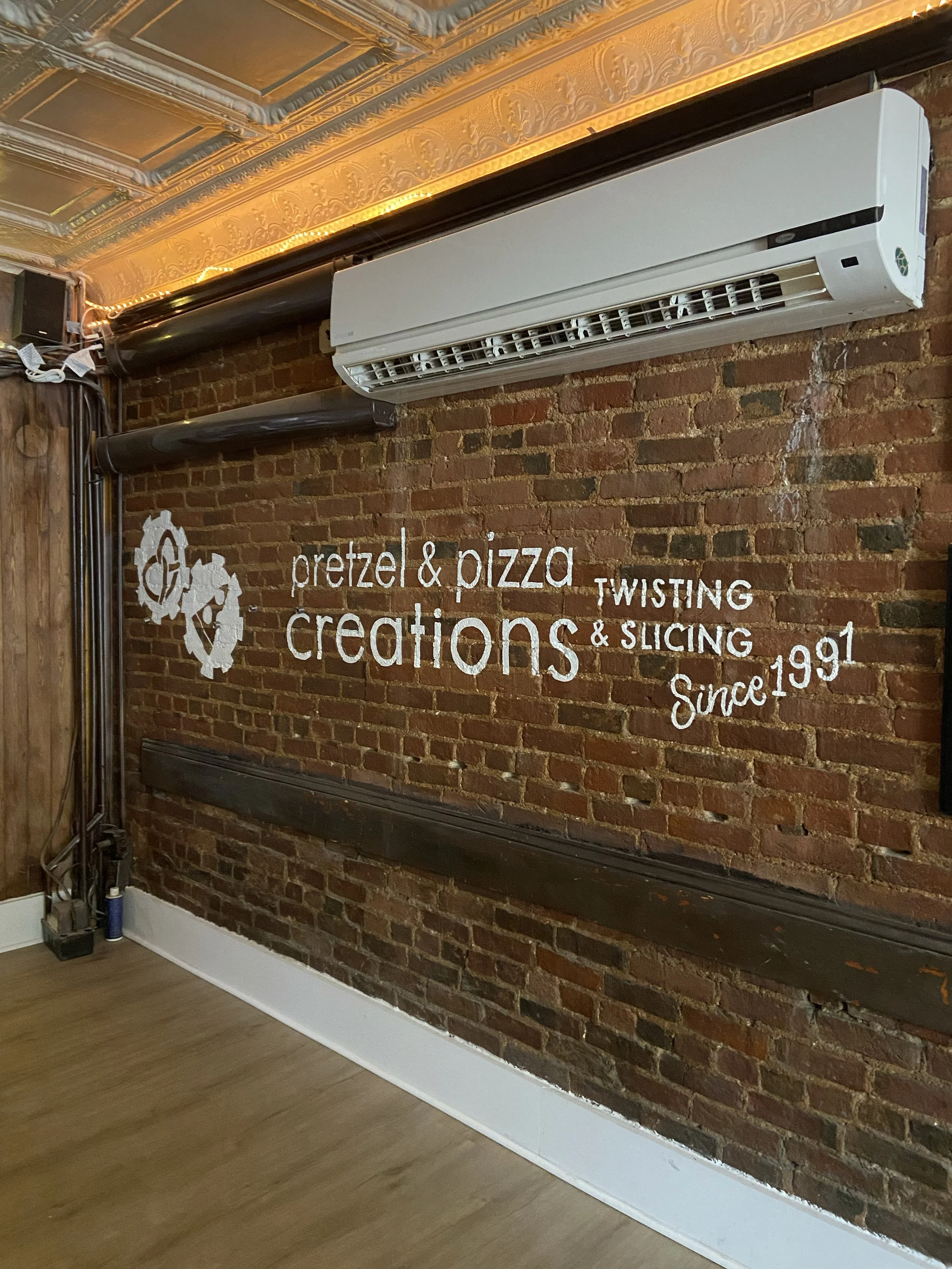Brick wall with white text and gears decor, advertising pretzel and pizza slice twisting, slicing, and creations since 1991
