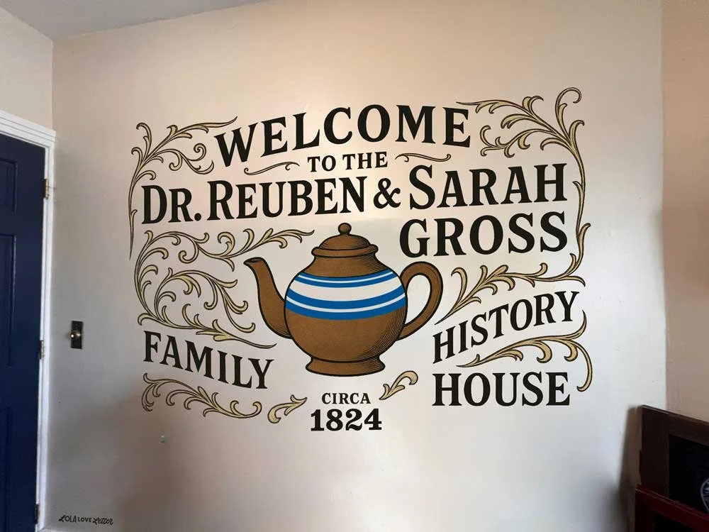 Wall with a hand painted welcome sign for the Gross family, mentioning family history from circa 1824, featuring a teapot illustration and ornate gold swirl designs.