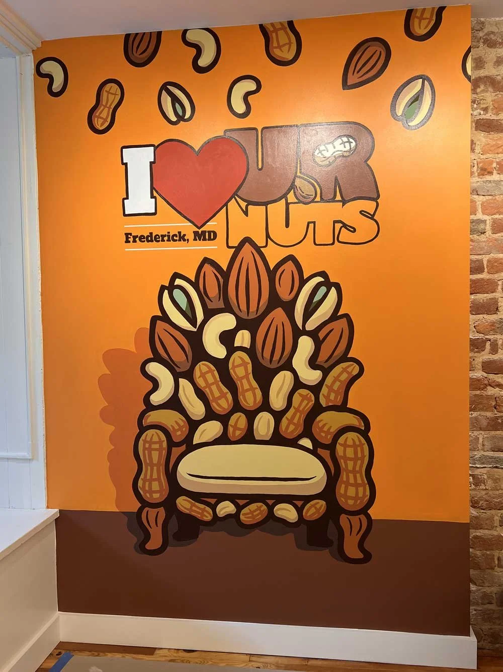 Bright orange wall with a cartoon illustration of a wooden chair filled with various nuts, with the text 'I love local nuts' and 'Frederick, MD' written above the chair, surrounded by scattered illustrated nuts.