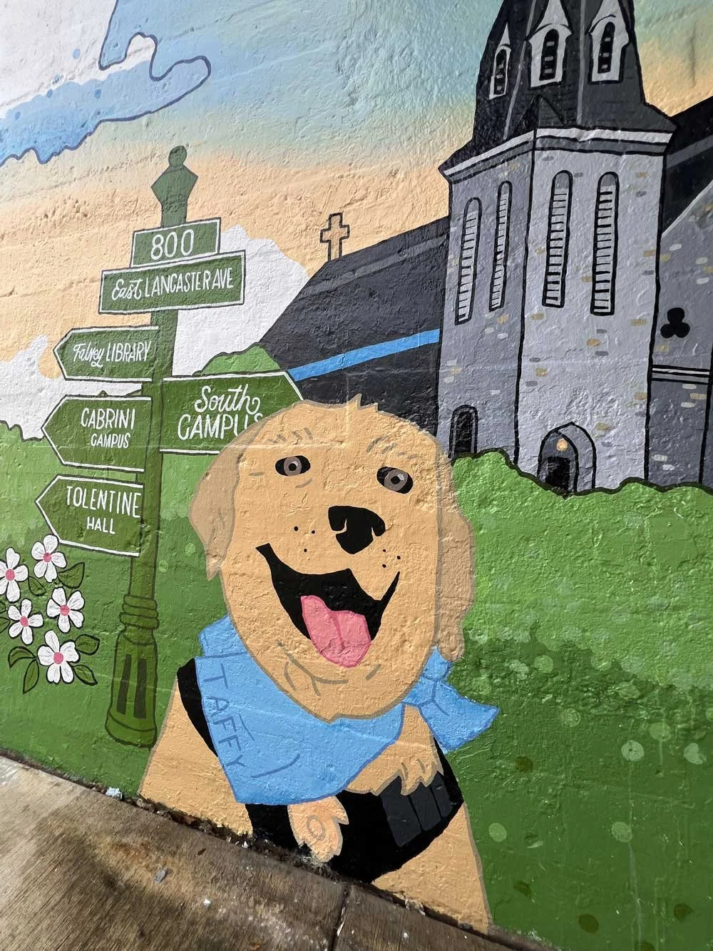 Colorful mural of a happy golden retriever dog with a blue bandana standing in front of a church and directional signpost in a cityscape scene.