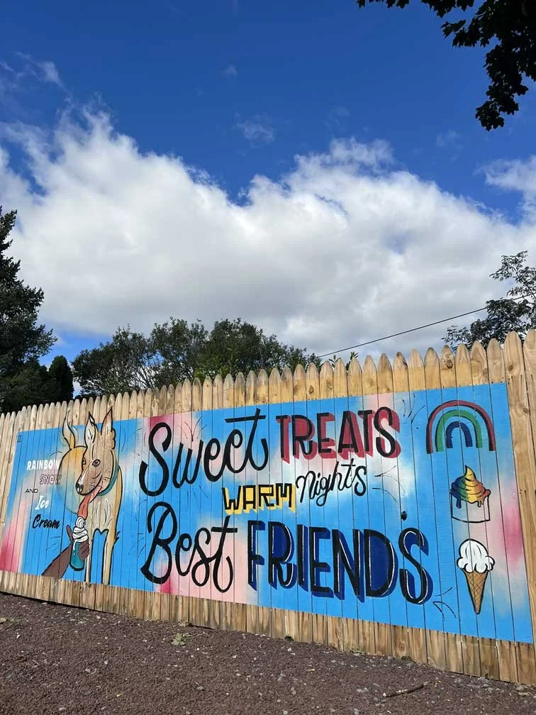 Wooden fence with a painted mural reading 'Sweet Treats Warm Nights Best Friends'. The mural features a dog eating a pup cup, a rainbow, an ice cream sundae, and an ice cream cone. The background shows a bright blue sky, partly cloudy day, and trees.