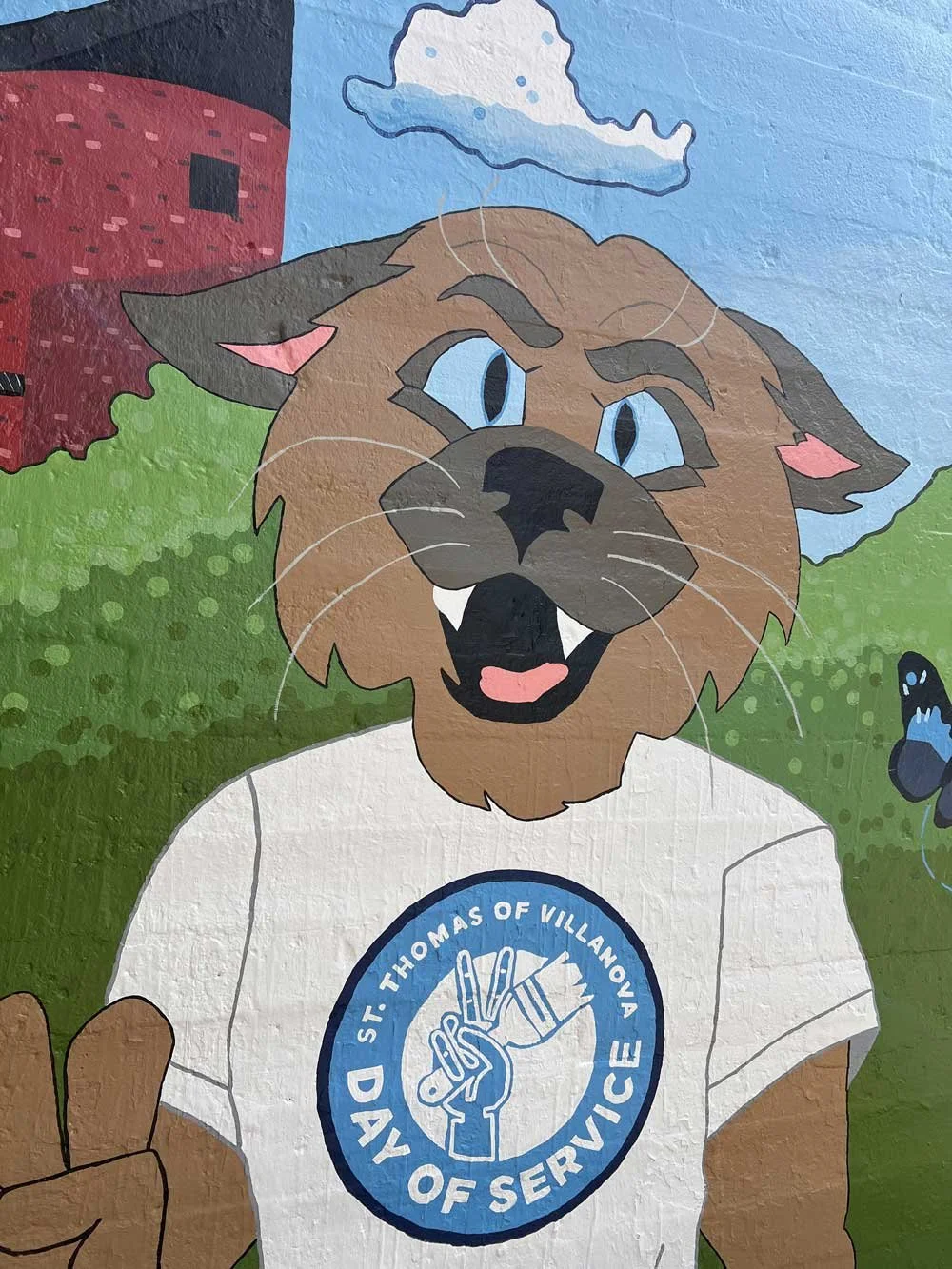 A colorful mural of a lion character wearing a white T-shirt with a 'St. Thomas of Villanova' logo, standing outdoors with greenery, a blue sky, a cloud, and a red brick building in the background.