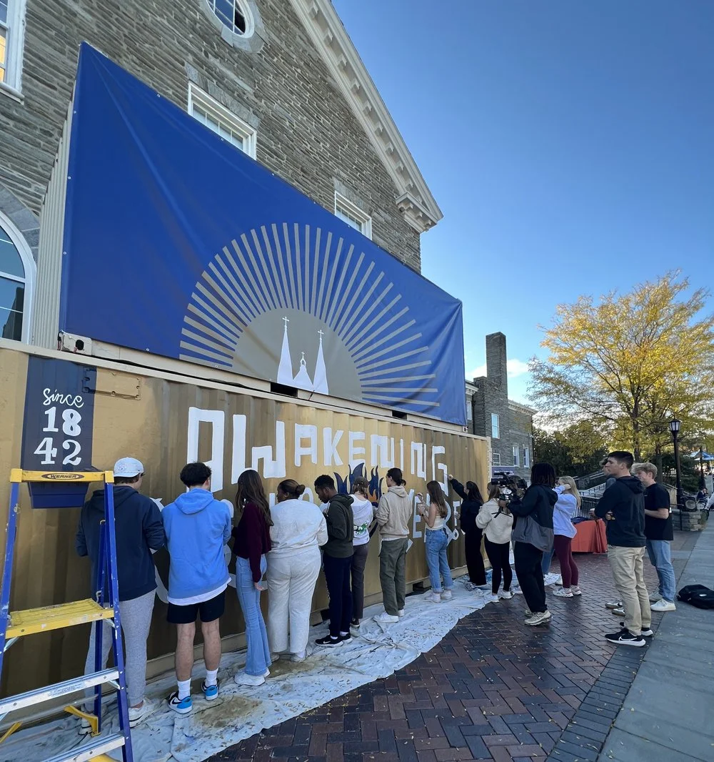 People standing in line outside a shipping container with a large banner above it, on a city street in the daytime. The banner displays a church steeples and rays of light. The container has the words 'PAWHAKENG GOLD' painted on it, and people are wa