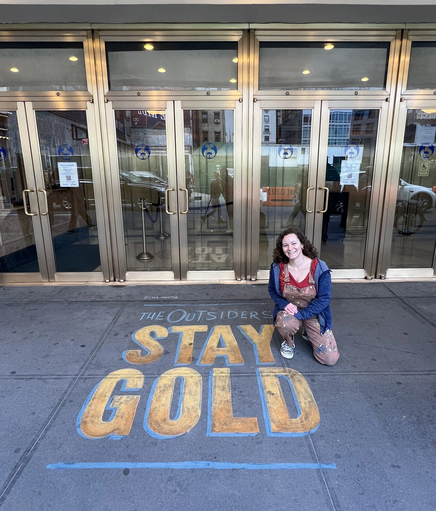 Lola Love Letter kneeling on a city sidewalk in front of a building with glass doors, smiling at the camera. On the sidewalk, there is a painted message that reads 'The Outsiders Stay Gold' in large yellow and blue letters.