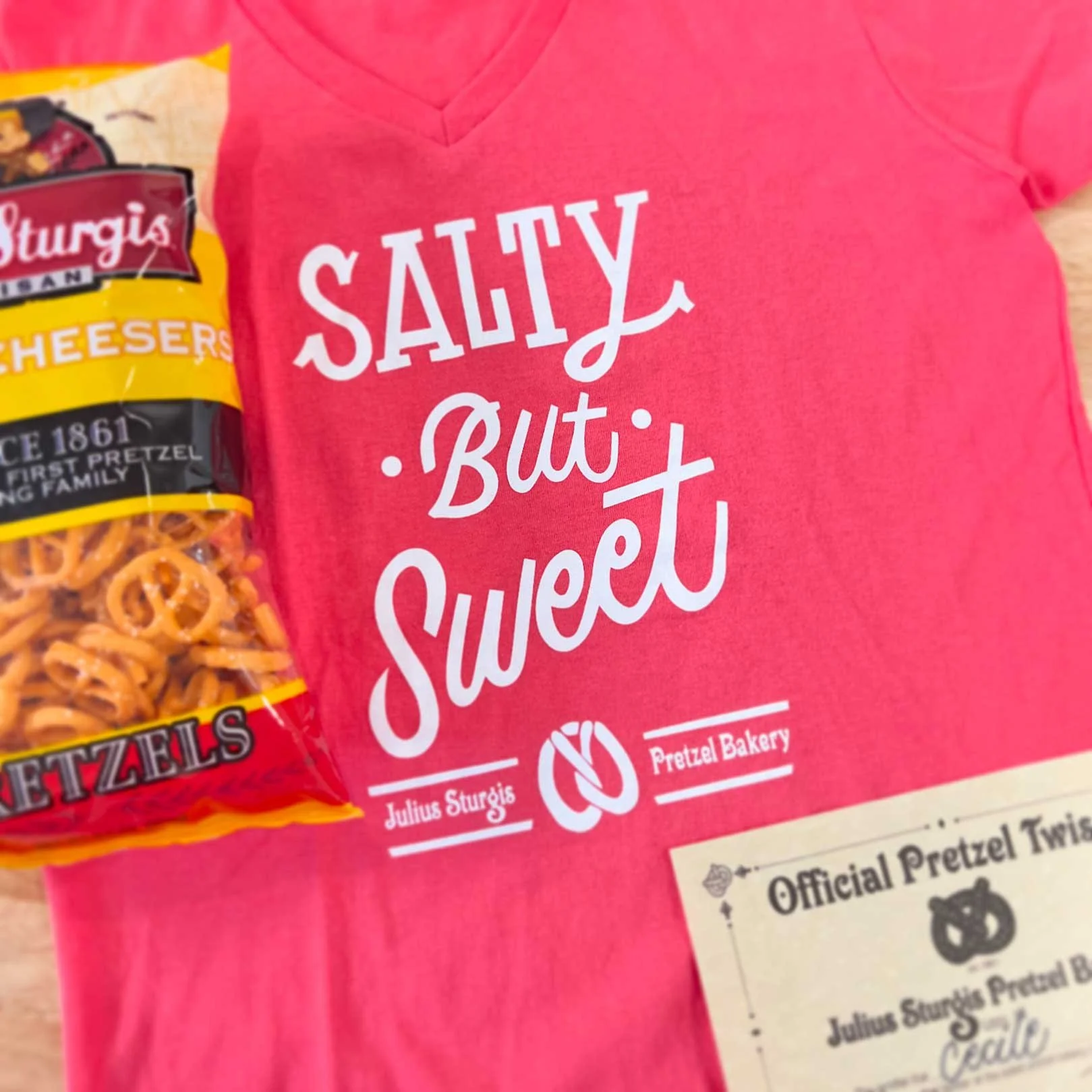 Pink t-shirt with white text saying 'Salty But Sweet' next to a bag of pretzels and a certificate from Julius Sturgis Pretzel Bakery.
