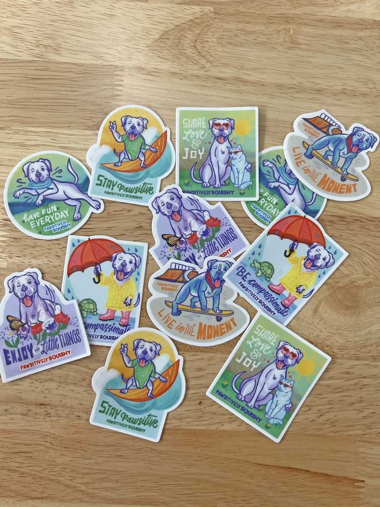 Colorful stickers featuring a cartoon dog in various playful and inspirational poses, with positive messages like "Stay Pawsitive," "Enjoy Little Things," "Share Love & Joy," and "Live in the Moment," on a wooden surface.