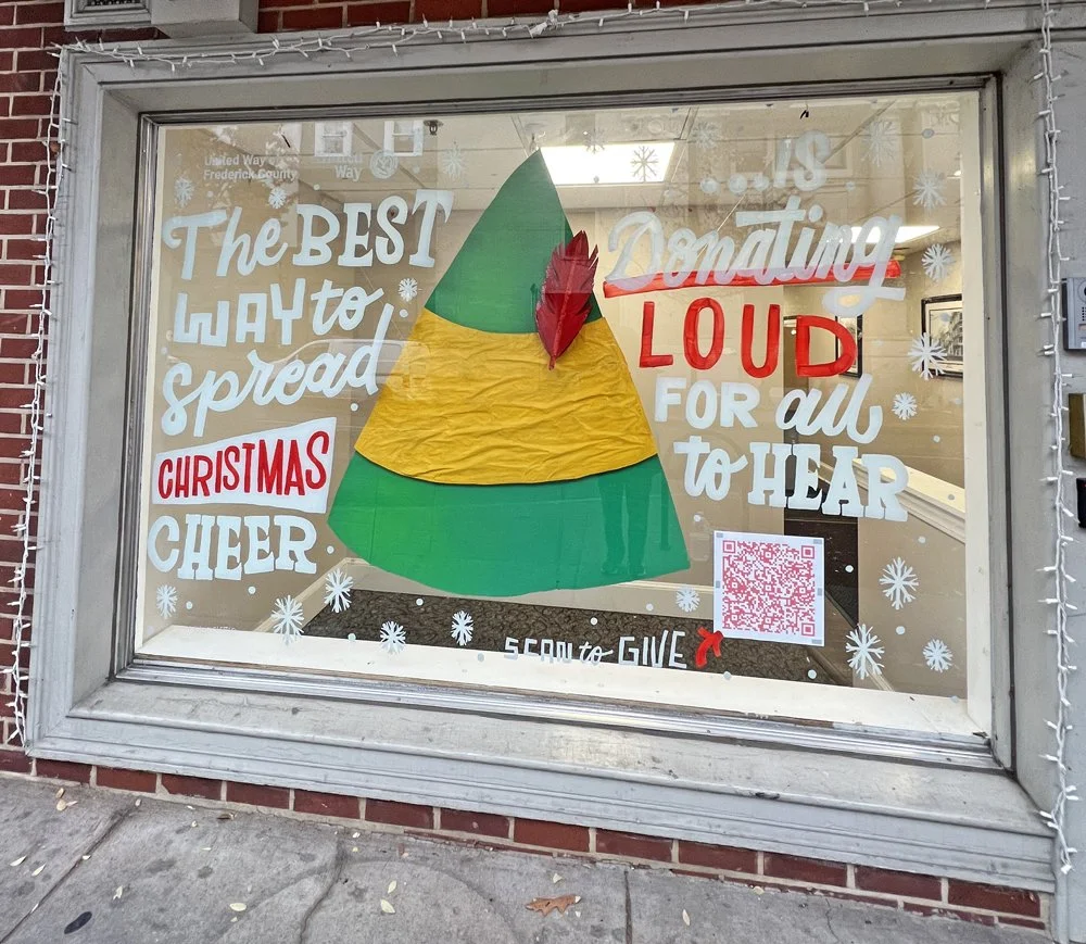 Store window decorated with Christmas message encouraging donations, with a paper Christmas tree and snowflake cutouts.