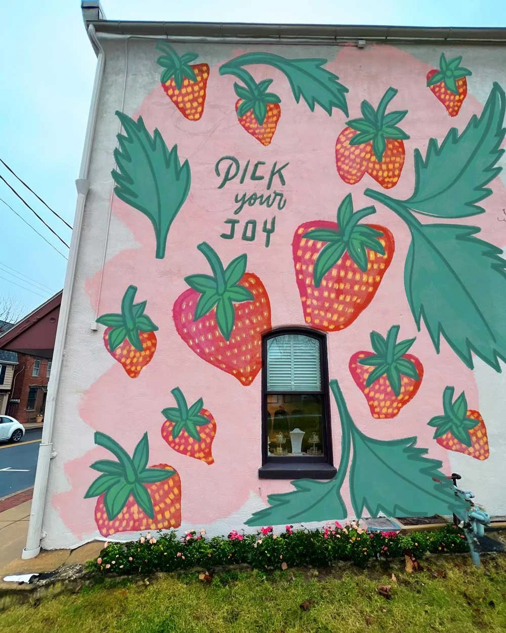 A mural painted on the side of a building featuring large strawberries with green leaves and the text "Pick your joy" in the center, set against a pink background. There is a small window at the bottom of the mural and some flowers planted beneath.