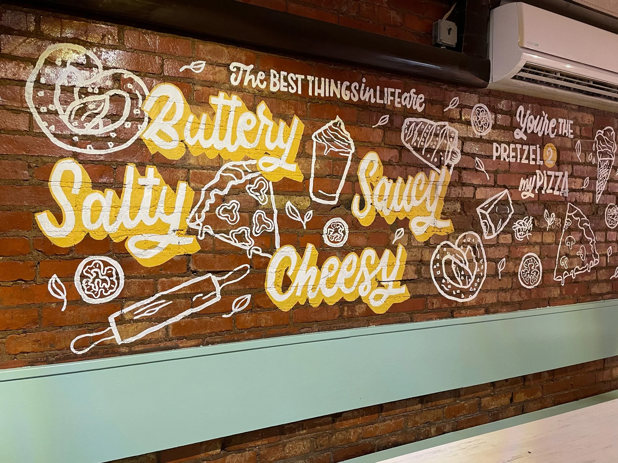 Decorative wall art with white and yellow illustrations of pizza slices, pretzels, cheese, and a drink, with phrases like 'The best things in life are buttery, salty, cheesy,' and 'you're the pretzel to my pizza,' on a brick wall background.