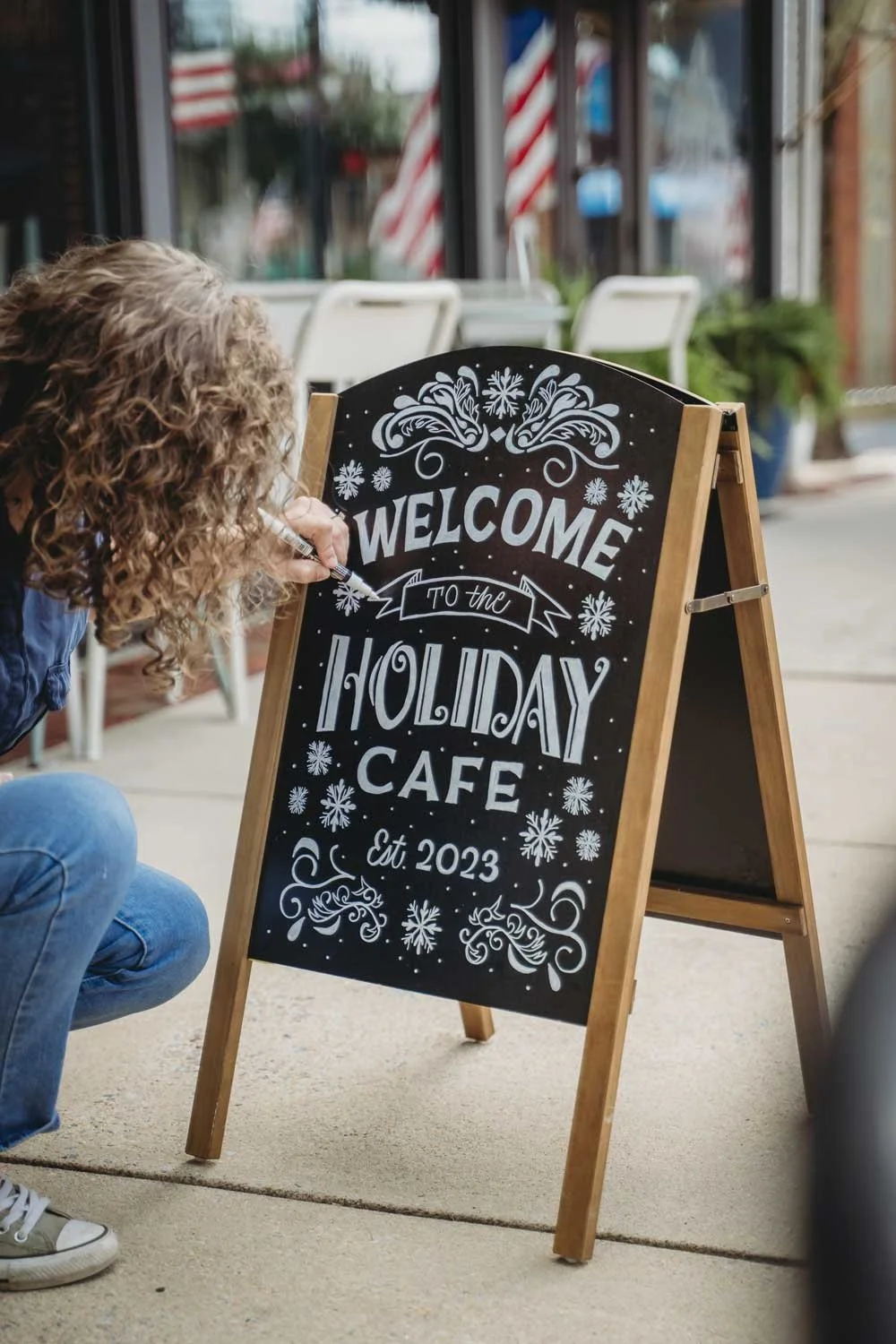 Lola Love Letter writing on a chalkboard sign outside a cafe. The sign reads, 'Welcome to the Holiday Cafe, Est. 2023,' with festive snowflake and swirl decorations.