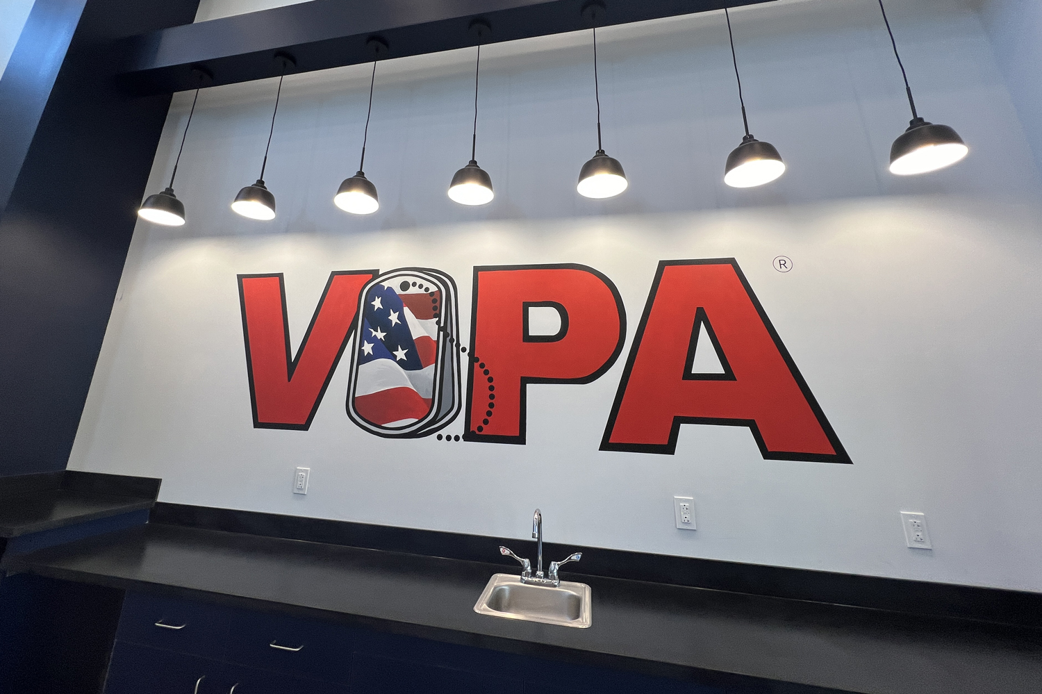 Wall with the word VOPA in large red letters, with an American flag pattern inside the letter 'O,' under six hanging lights, a small sink, and electrical outlets on the wall.