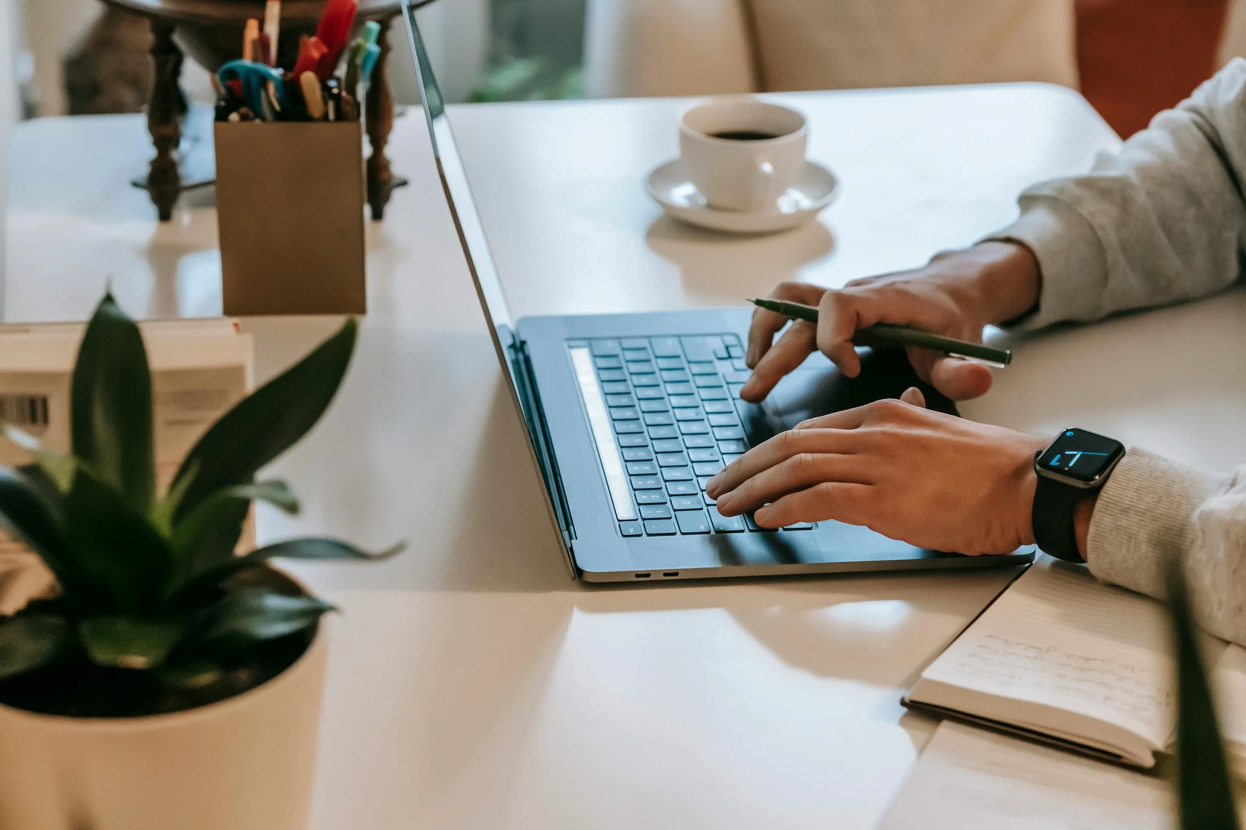 Person typing on a laptop at a desk with a coffee cup, notebook, plant, and pen holder.