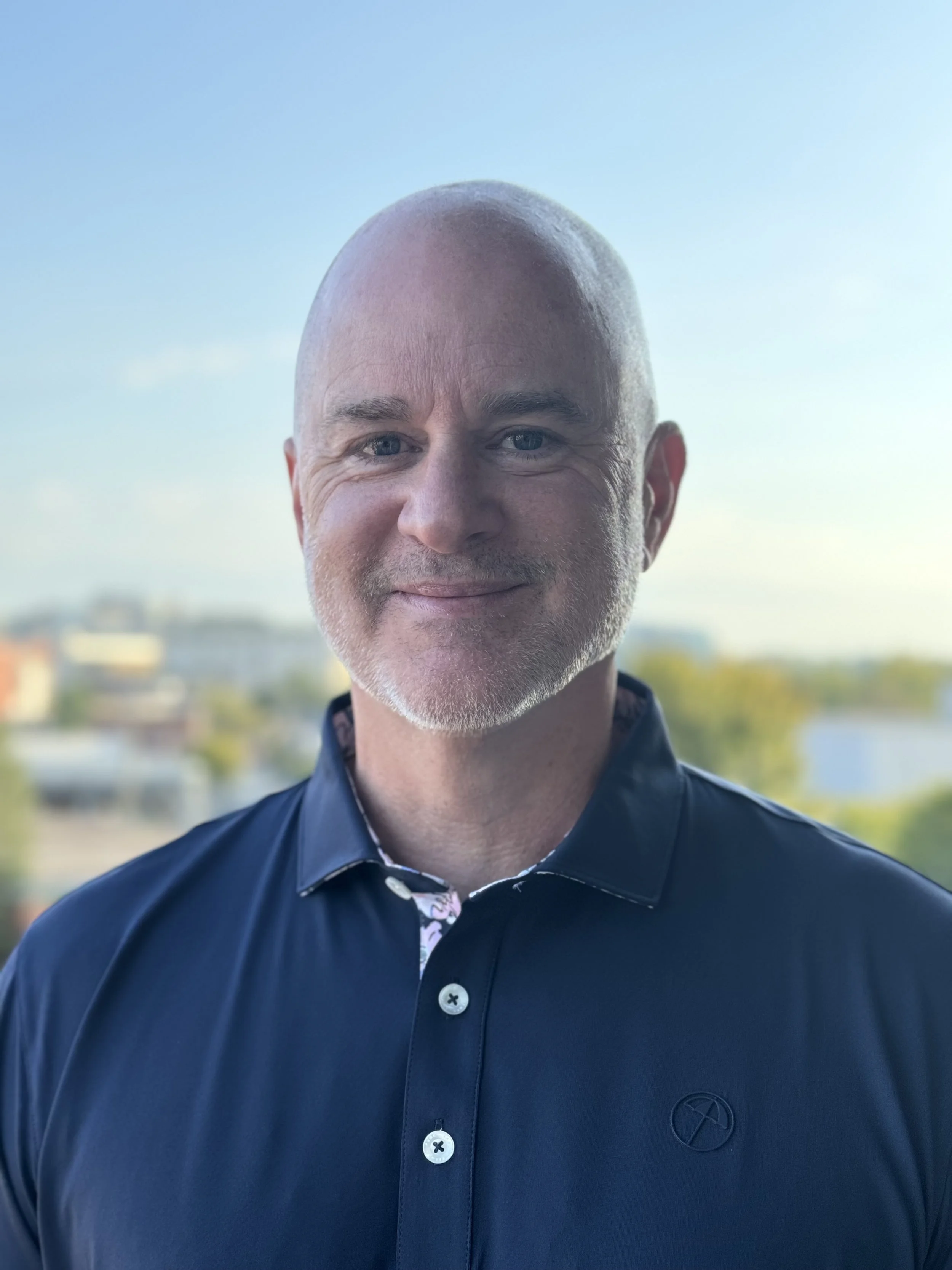 Bald man in dark shirt smiling against blurred outdoor background