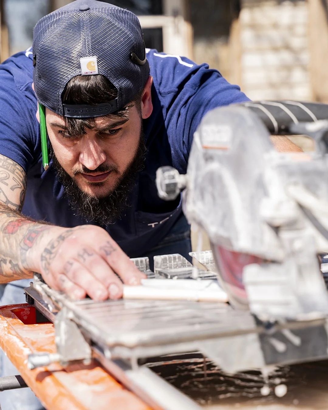 Man wearing a baseball cap operating a wet tile saw, focusing intently on cutting a tile.