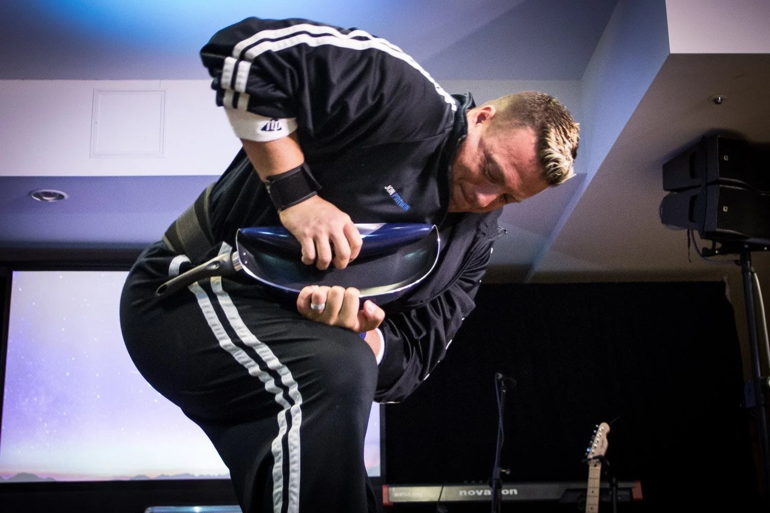 A person bending a metal object in a performance setting, wearing a black tracksuit.
