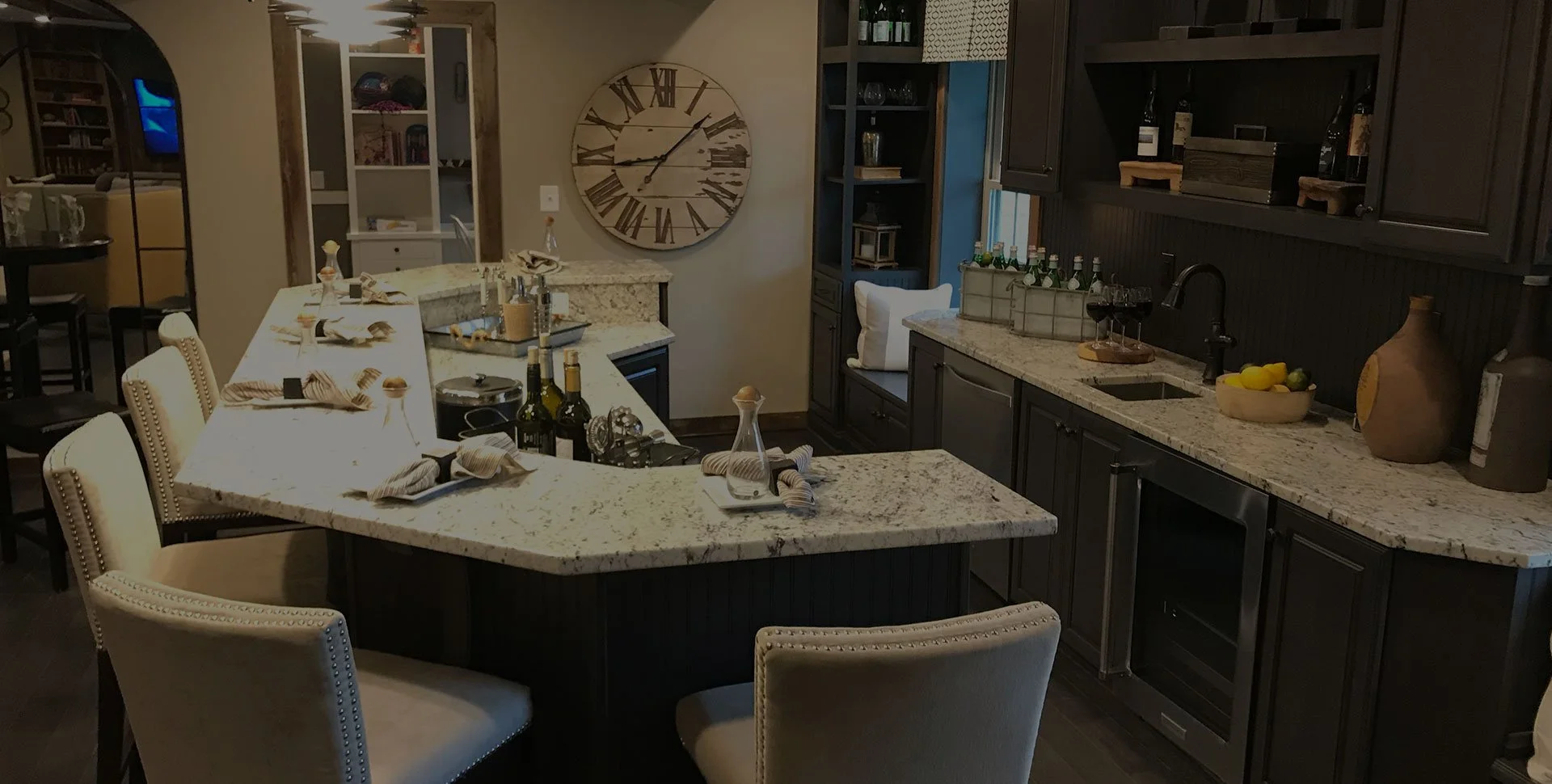 A kitchen with a granite island in the center, decorated with wine bottles and tableware, surrounded by beige chairs. Dark cabinets line the walls with a window seat underneath. Decor items include a large wall clock, pottery, bottles, fruit, and a w