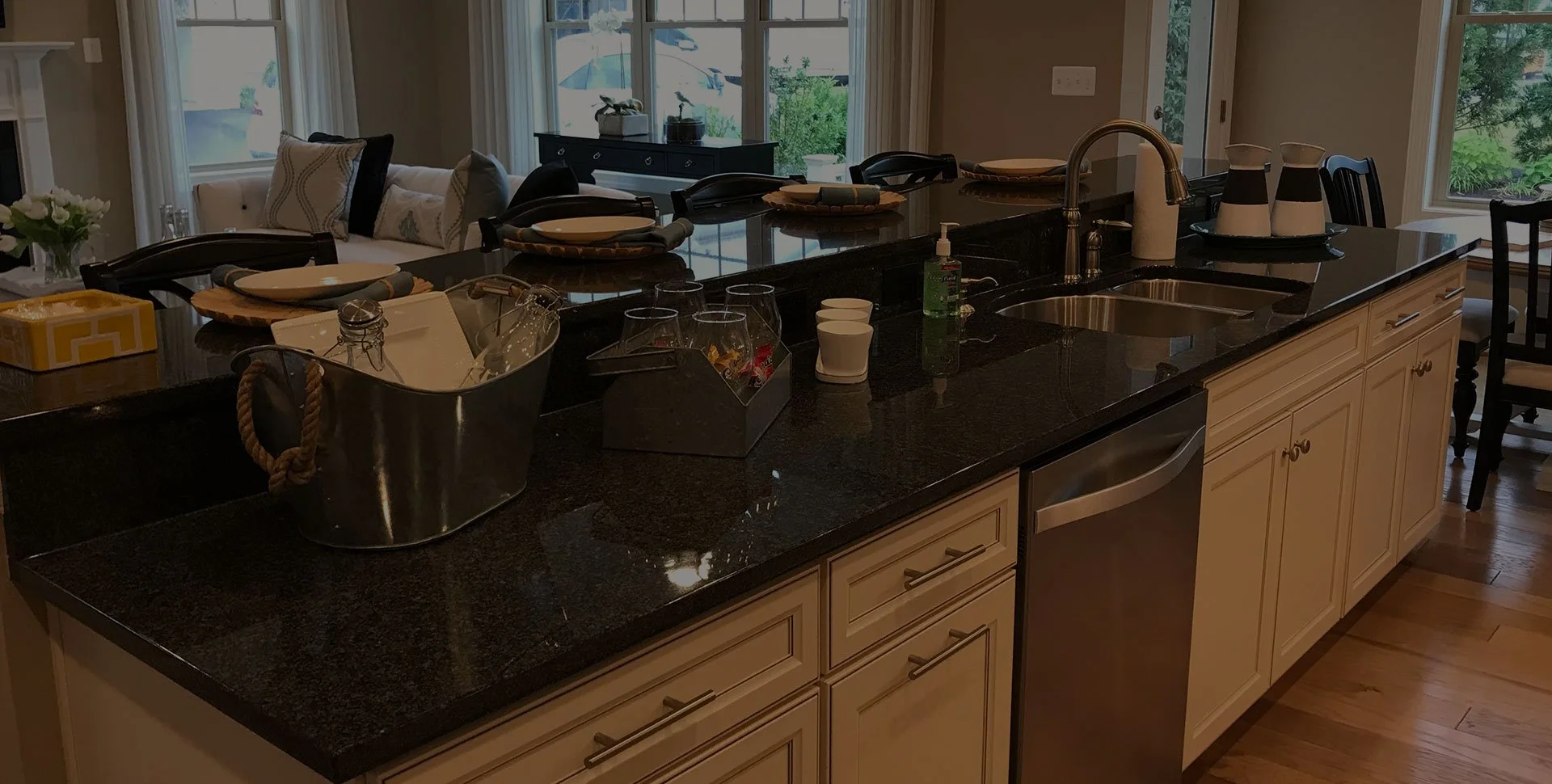 Kitchen island with black countertop, sink, and various items including a soap dispenser, cups, and a container. In the background, there is a living room with large windows, a sofa with pillows, and a dining area with chairs.