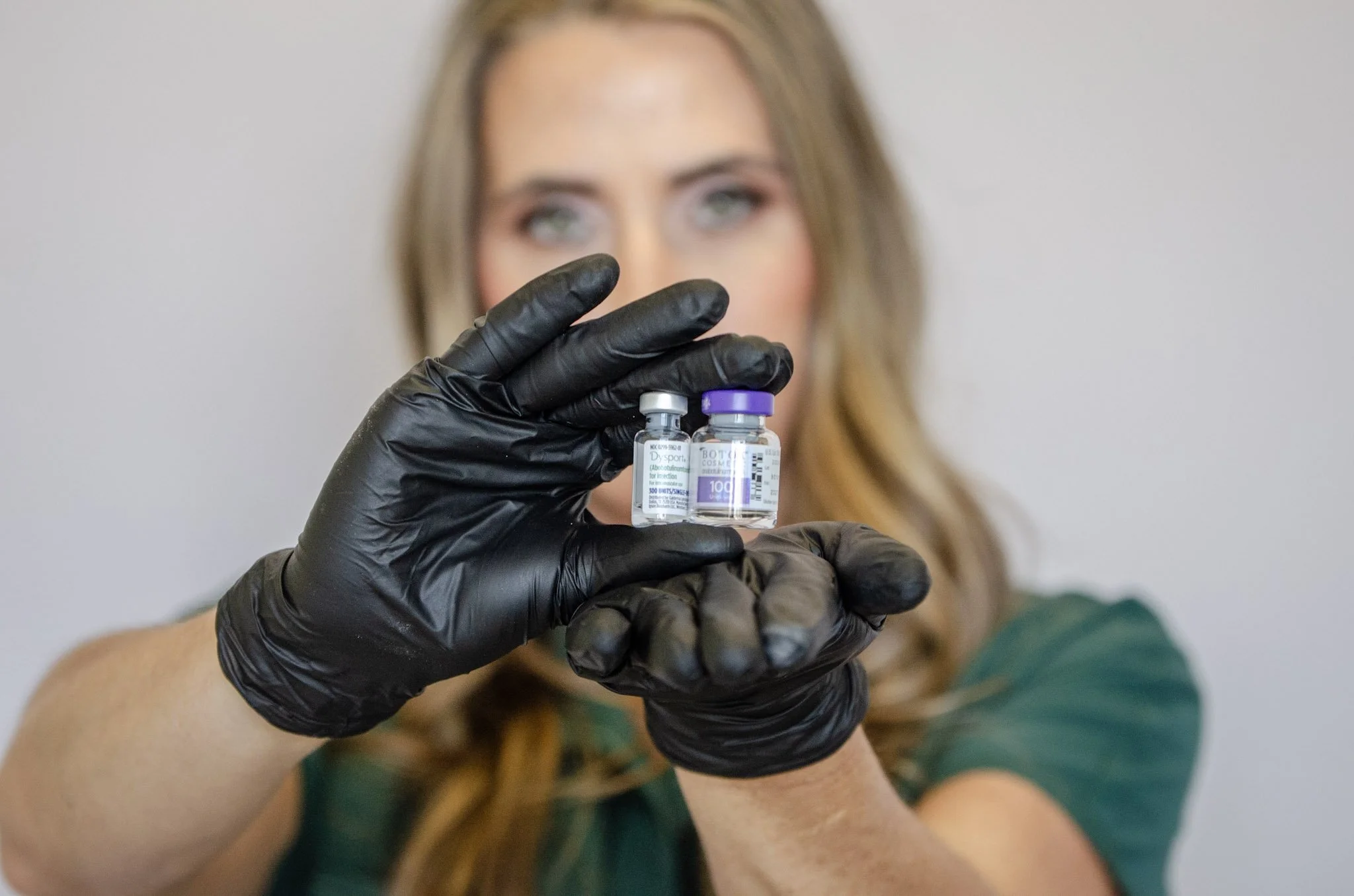 A woman with long hair wearing black gloves holding two small vaccine vials, with a blurred background.