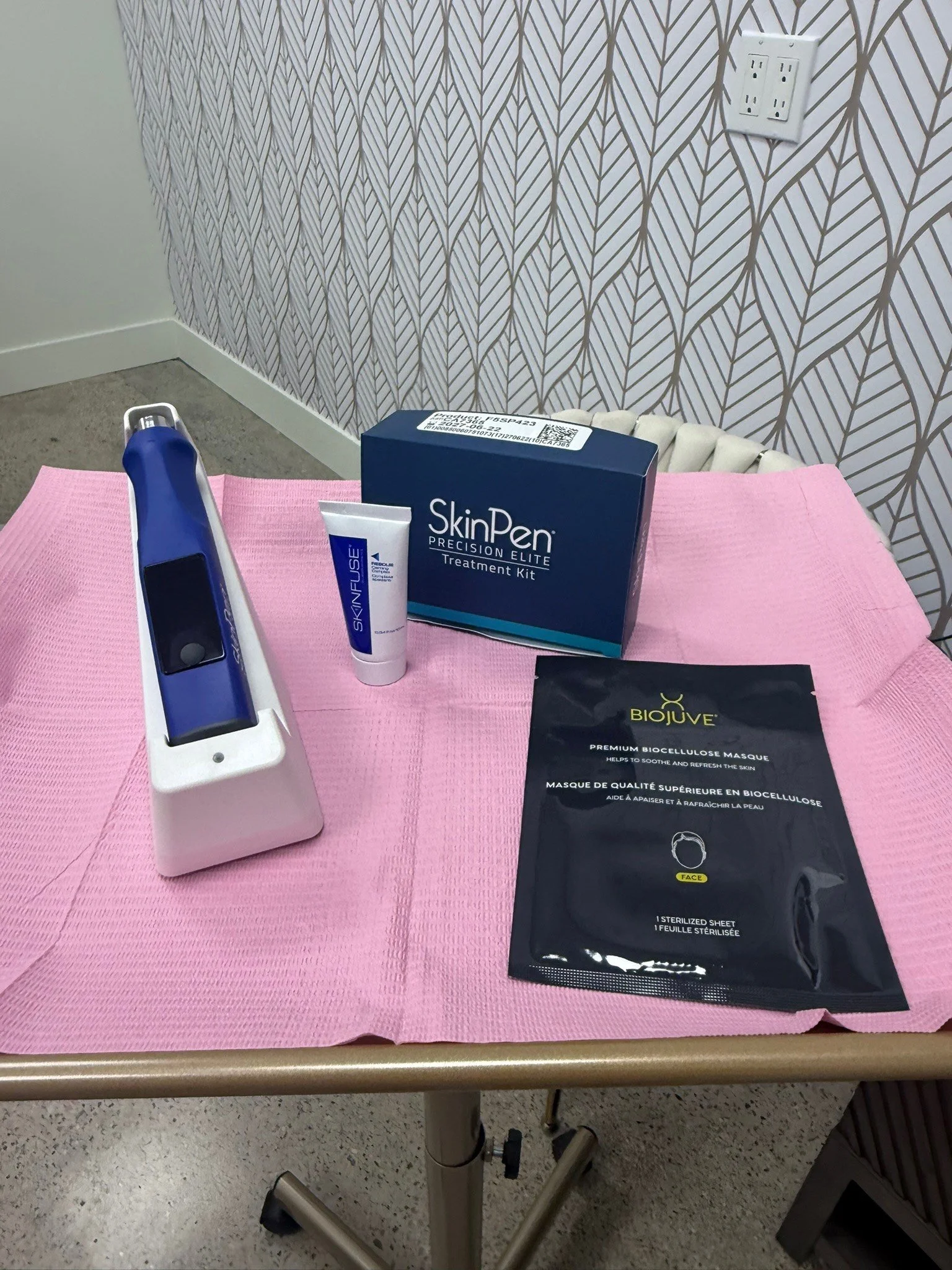 Medical treatment or skincare setup with a SkinPen Precision Elite treatment kit, a tube of SkinFuse, a bio-cellulose face mask, a sterilized sheet, and a blue device on a pink sheet on a medical table.