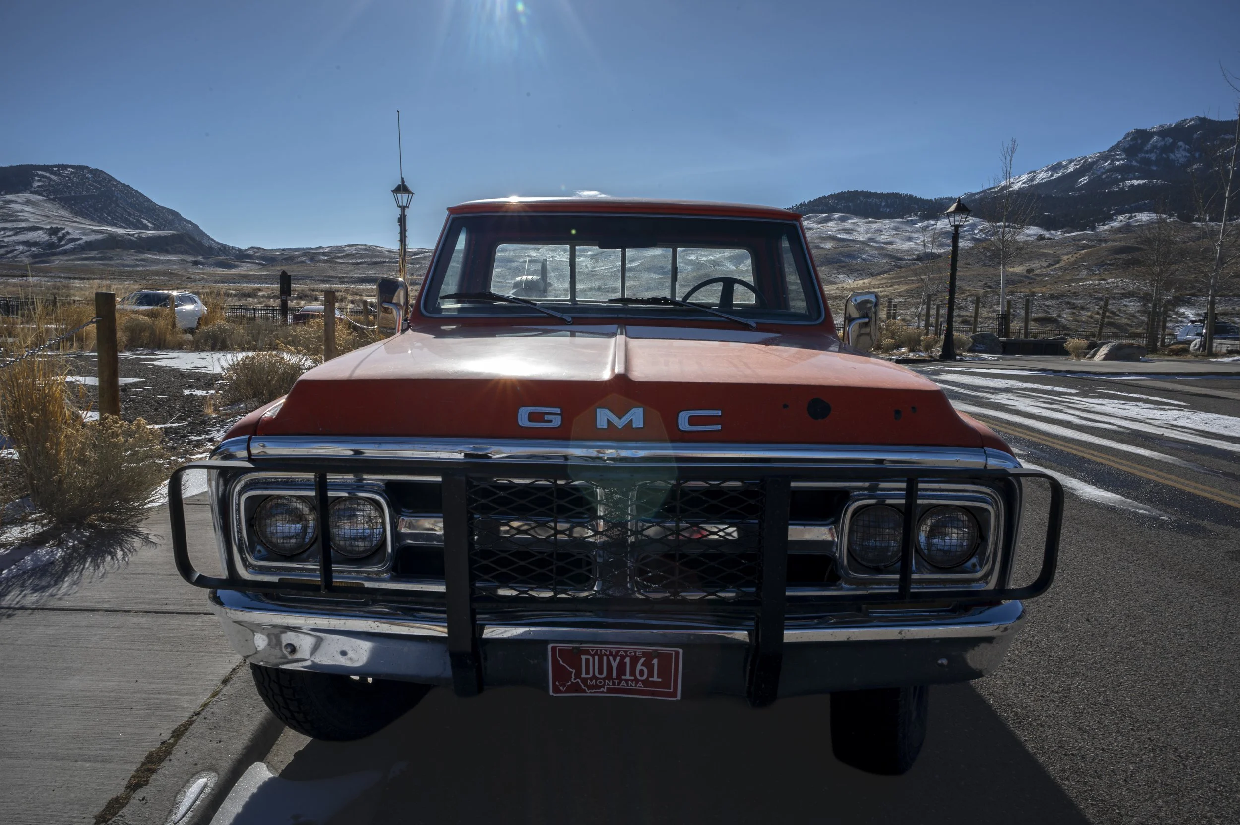 Front view of a vintage red GMC truck with a black grille guard on a snowy mountain road under a clear blue sky.