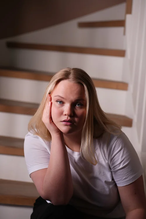 Ung kvinne med blondt hår, sitter på stige med hvit t-skjorte og ser direkte i kamera, med hodet støttet av hånden.