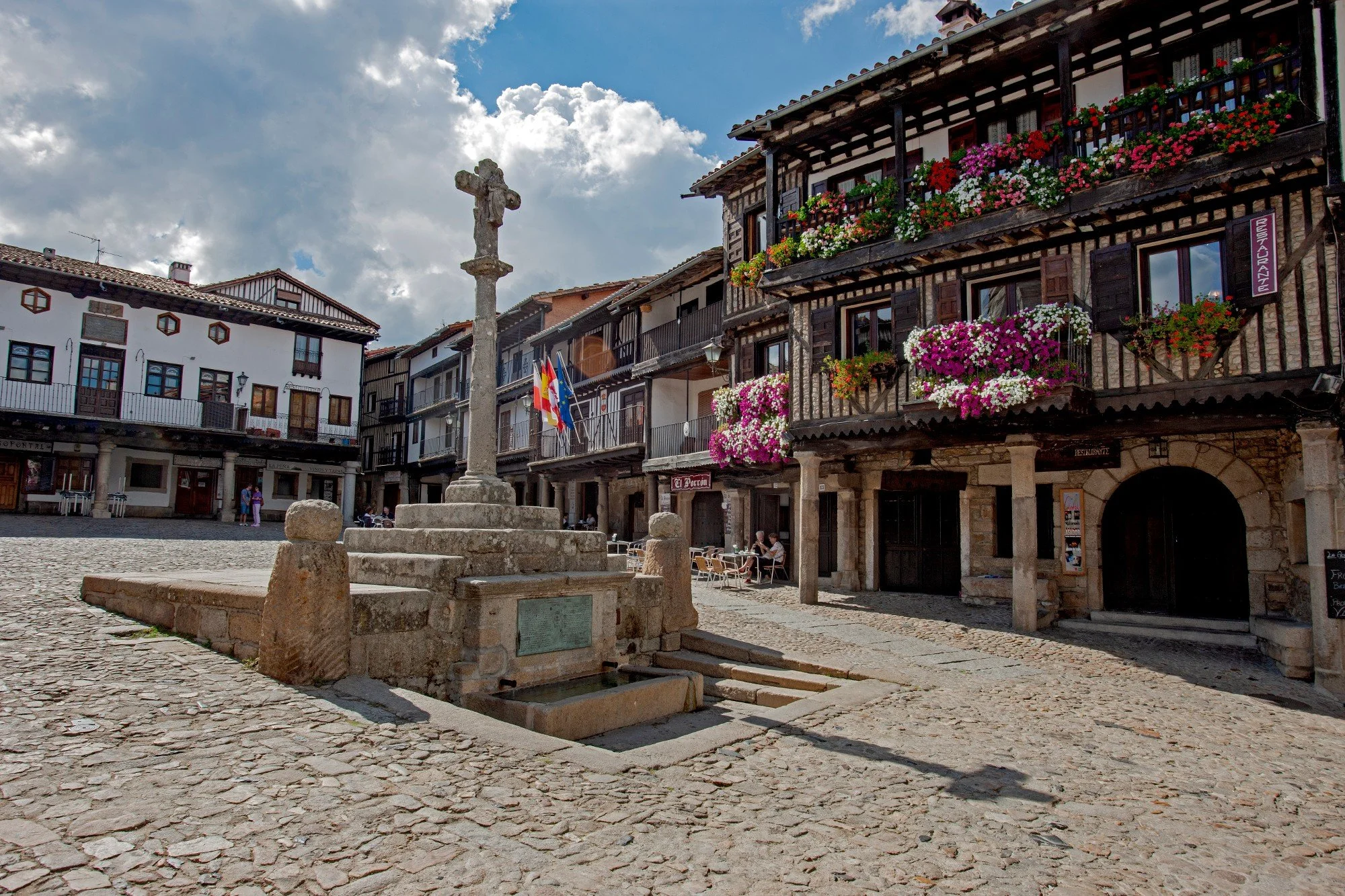 Plaza de piedra con un monumento en el centro y edificios tradicionales de madera y piedra con flores en los balcones. Hay un par de personas sentadas en mesas, y banderas en la fachada.