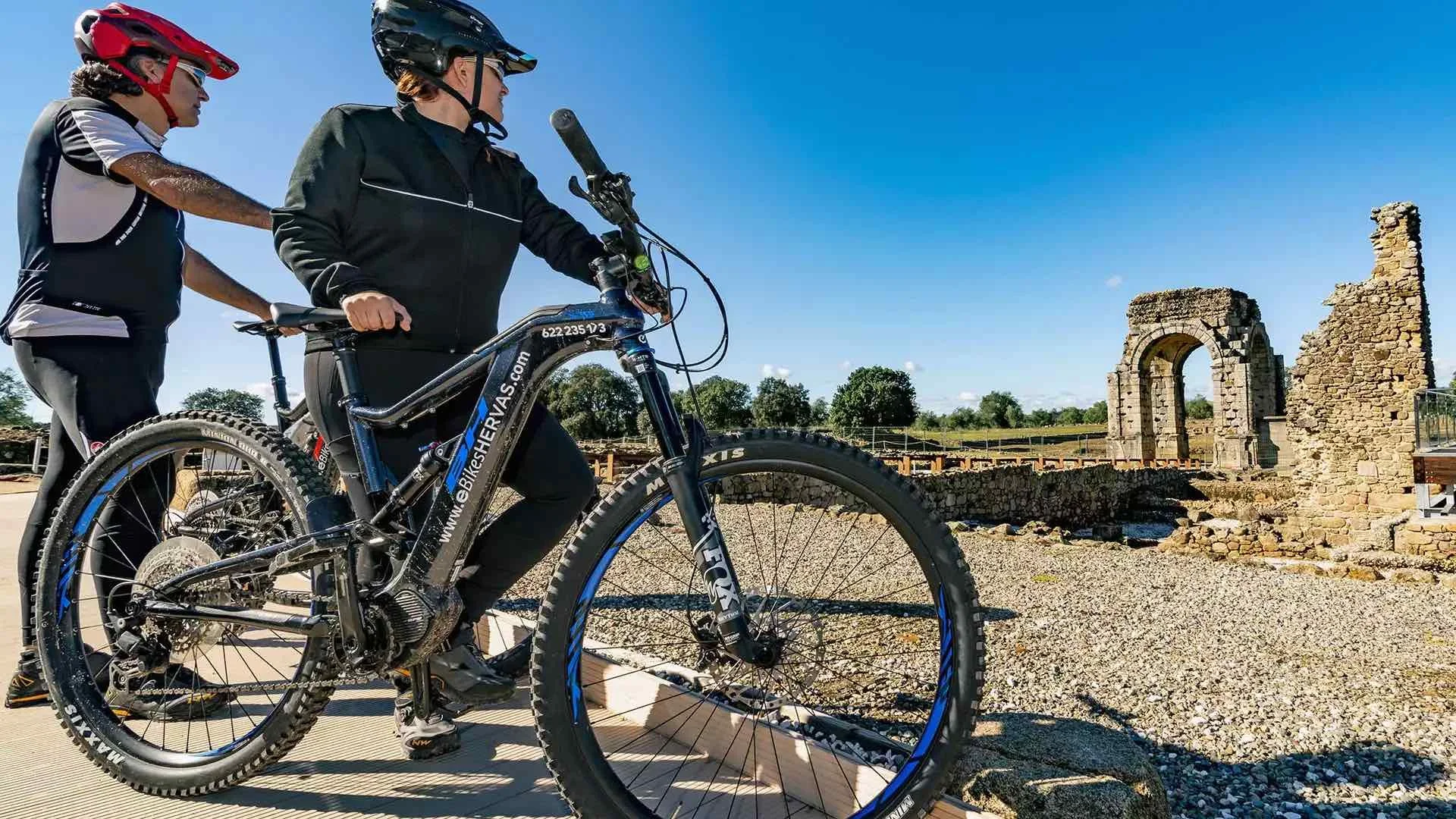 Dos personas en bicicleta de montaña en un sitio arqueológico con ruinas de piedra y un arco, bajo un cielo despejado.