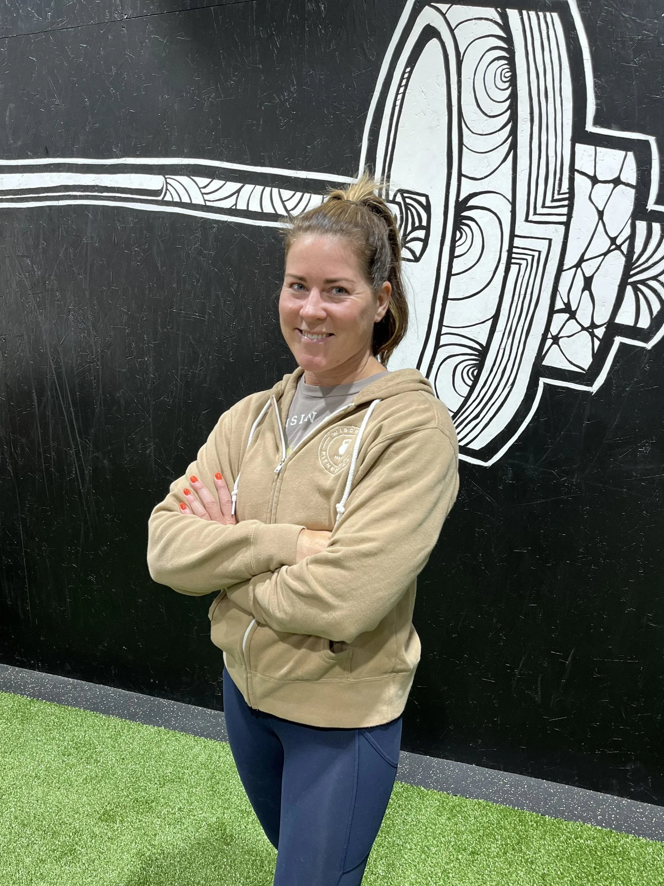 Woman with brown hair in a ponytail, wearing a beige zip-up hoodie and navy blue leggings, standing with arms crossed in front of a black wall with a white graphic mural.