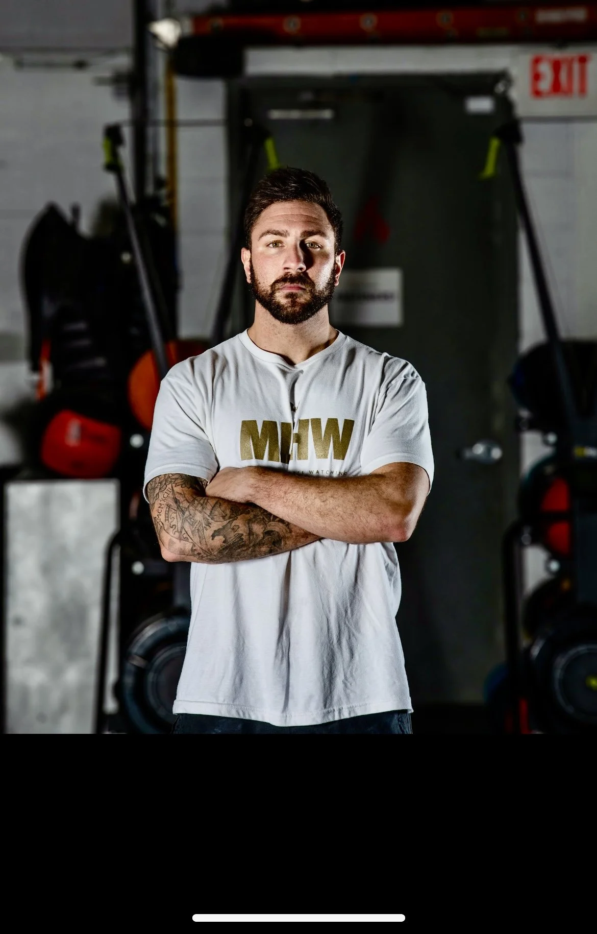 A man with a beard, dark hair, and tattoos on his left arm, standing with crossed arms in a gym. He's wearing a white T-shirt with gold lettering that reads 'MEW'. The gym background has various equipment and an exit sign.