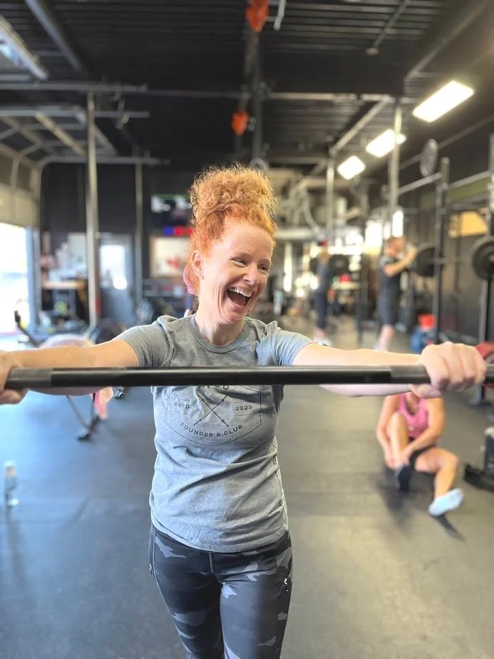 A woman with curly red hair smiling happily while holding a barbell in a gym. She is wearing a gray T-shirt and camo-patterned leggings. Other gym equipment and people are visible in the background.