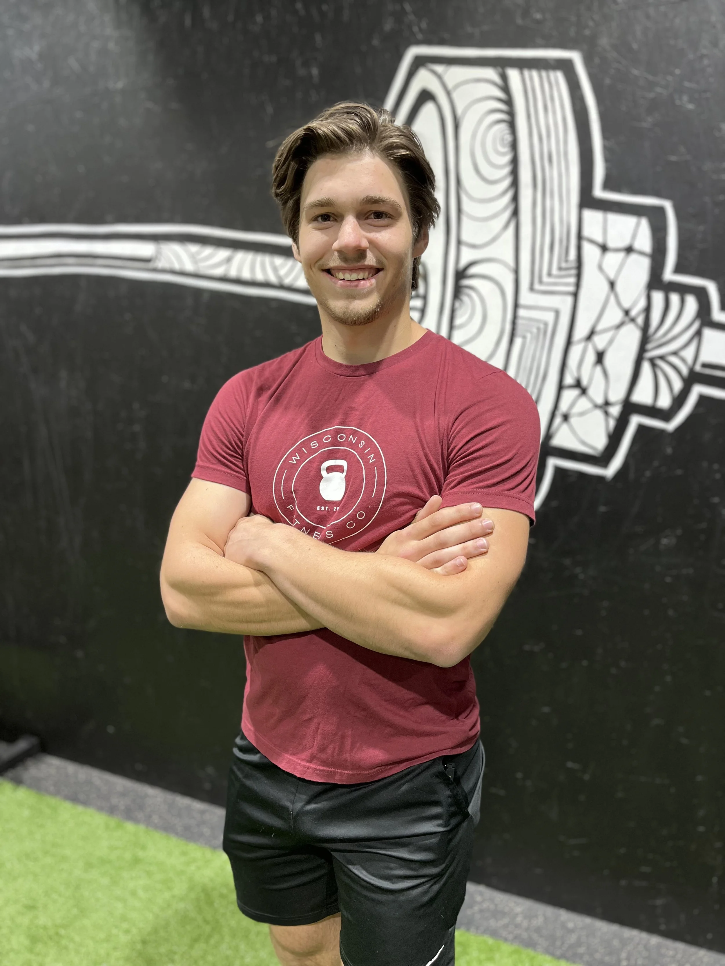 A young man with brown hair and a goatee smiling and standing with arms crossed in front of a black wall with white graffiti art, wearing a red T-shirt and black shorts.