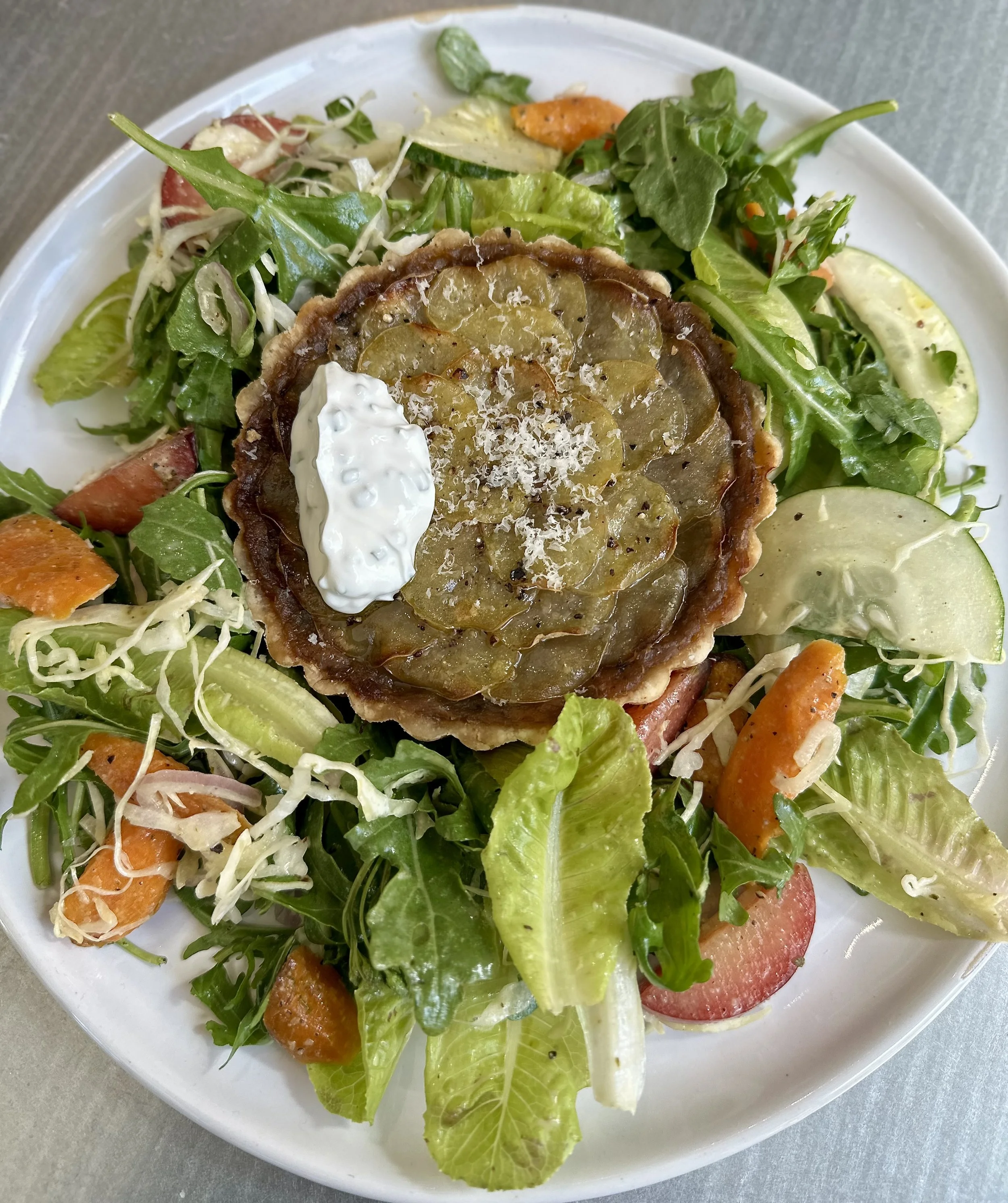 Salad with mixed greens, cherry tomatoes, slices of cucumber, shredded cheese, and a vegetable tart topped with sliced pickles and a dollop of creamy dressing on a white plate.