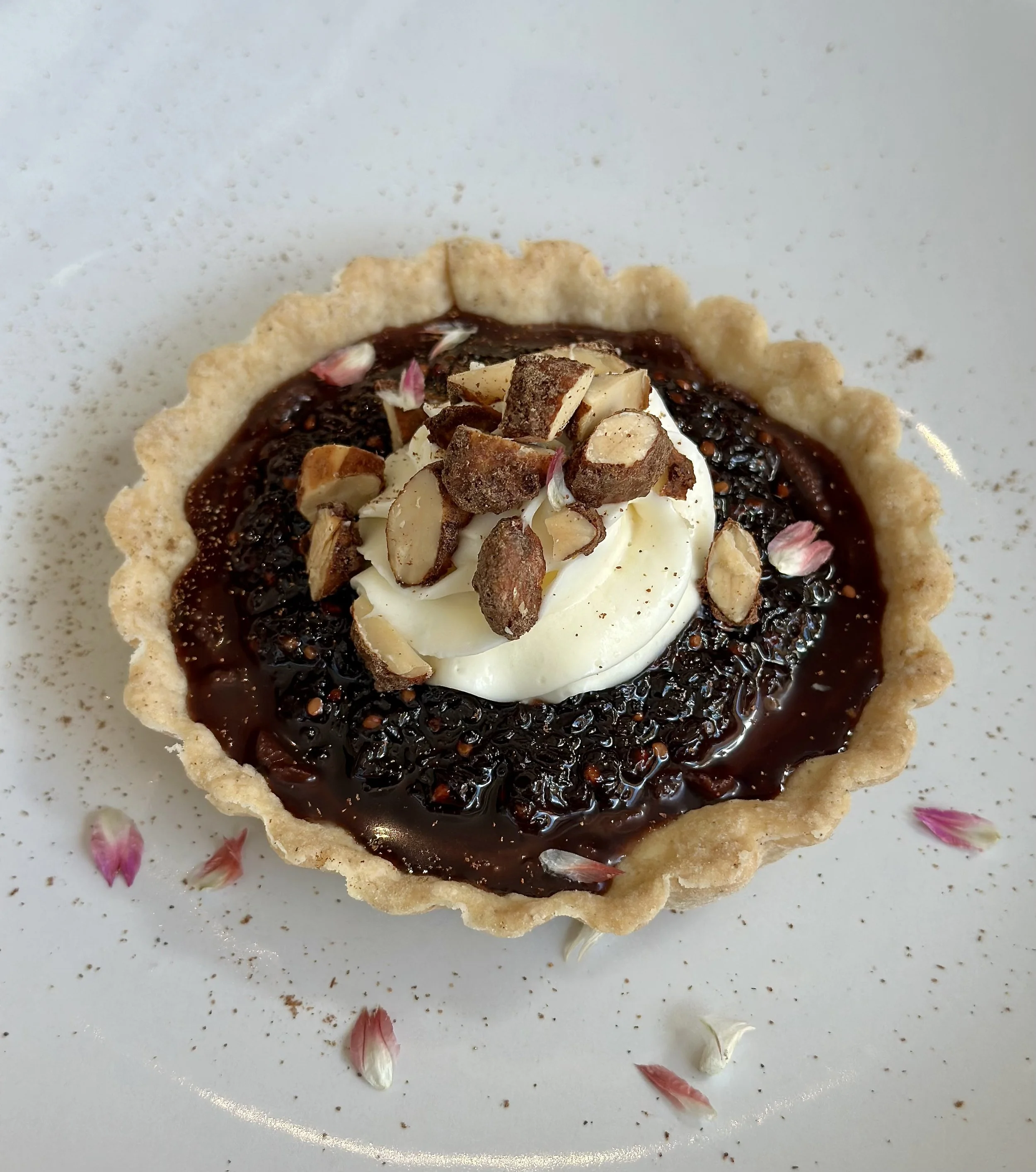 Chocolate tart garnished with whipped cream and chopped almonds, with pink and white flower petals on a white plate.