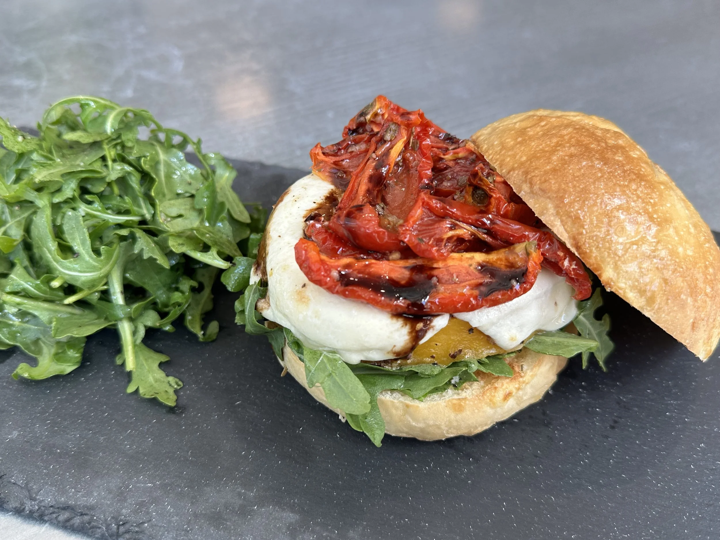 Close-up of a gourmet sandwich with roasted red peppers, melted cheese, greens, and a toasted bun, served with a side of fresh arugula on a black slate platter.