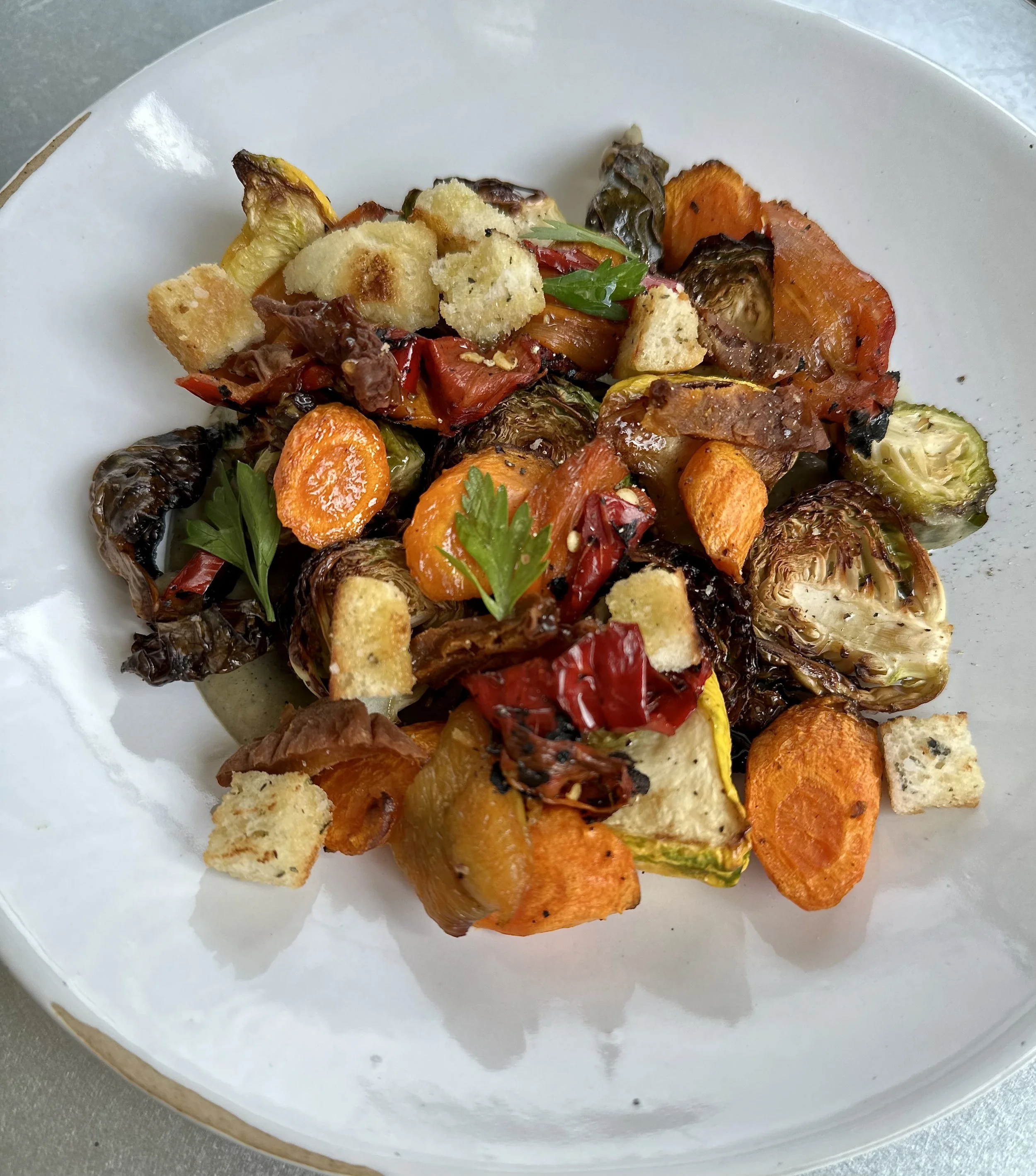 A plate of roasted mixed vegetables including carrots, Brussels sprouts, red and yellow bell peppers, and croutons garnished with fresh herbs.